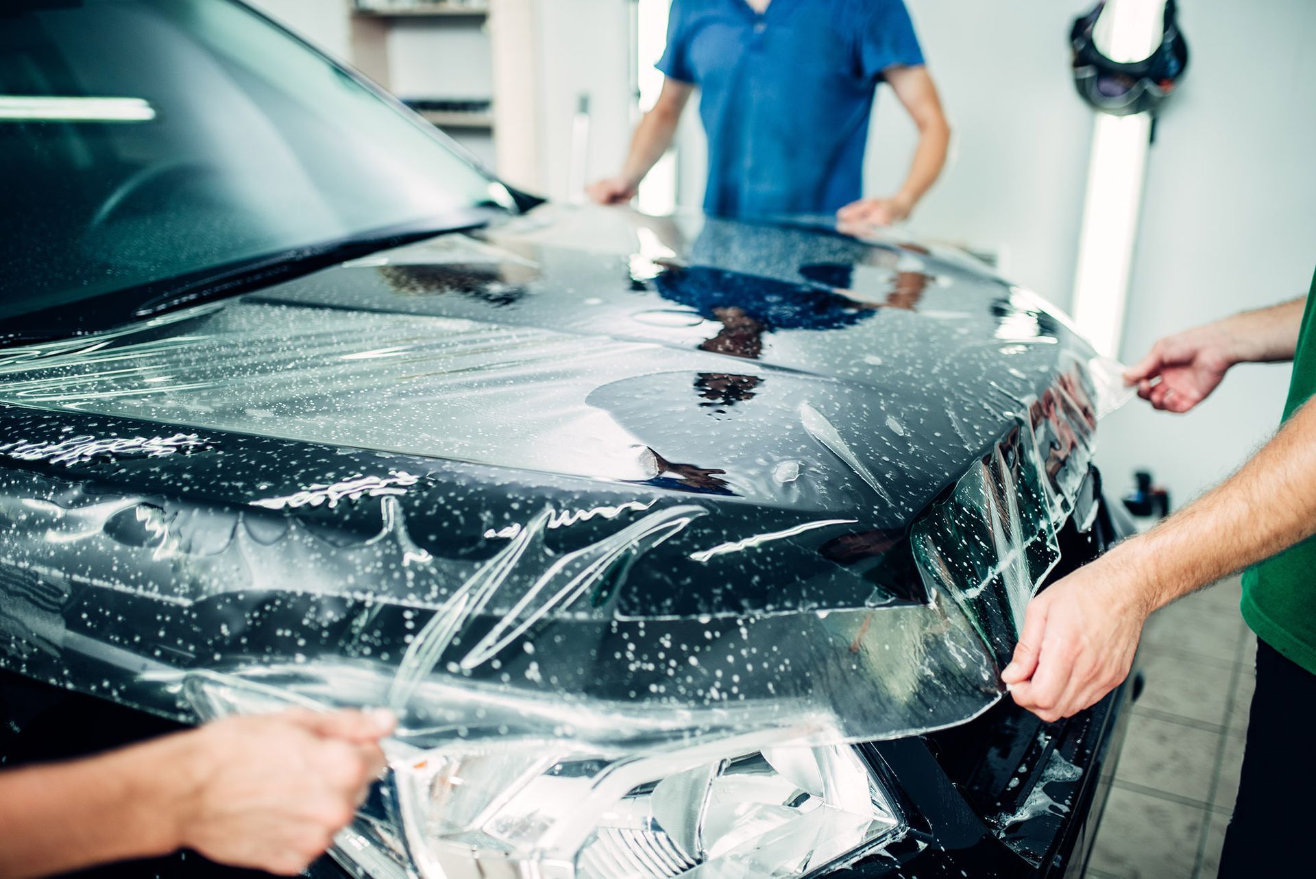 Two workers apply a protective transparent film onto the black hood of a car in a workshop.