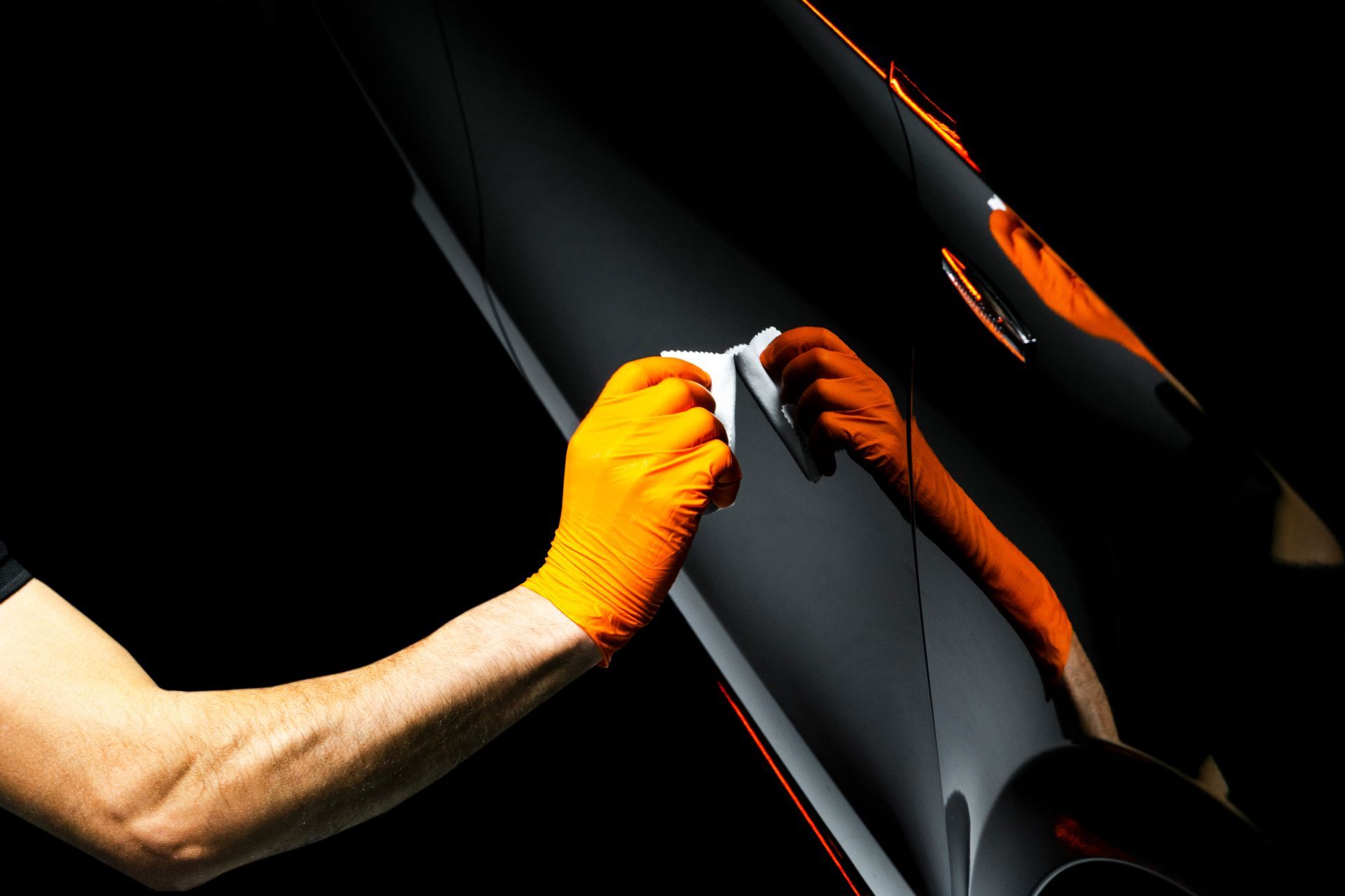A person wearing an orange glove wipes a black surface with a small white cloth against a dark background.