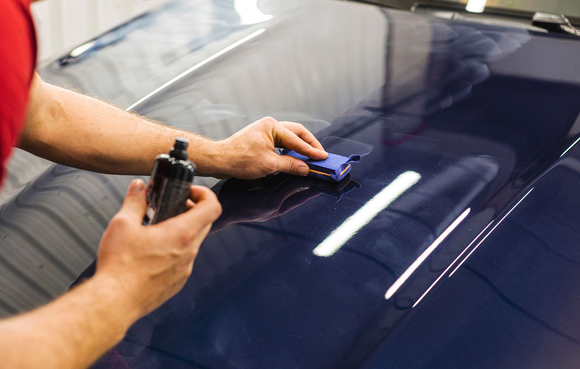 A person applies a ceramic coating to a dark blue car hood using a small applicator pad and a bottle of solution.