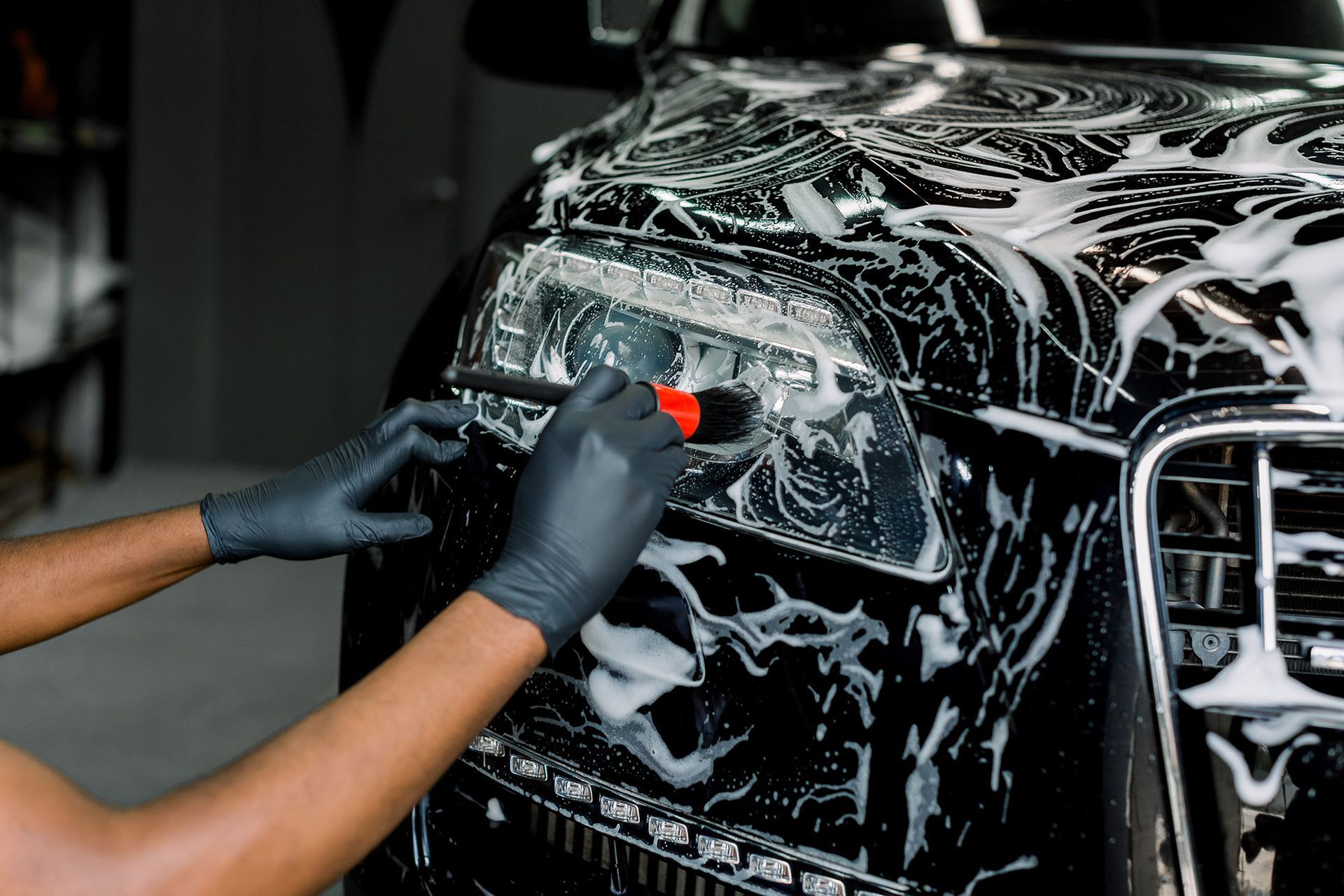 A person wearing black gloves uses a detail brush to clean the headlight of a black car covered in soap suds.