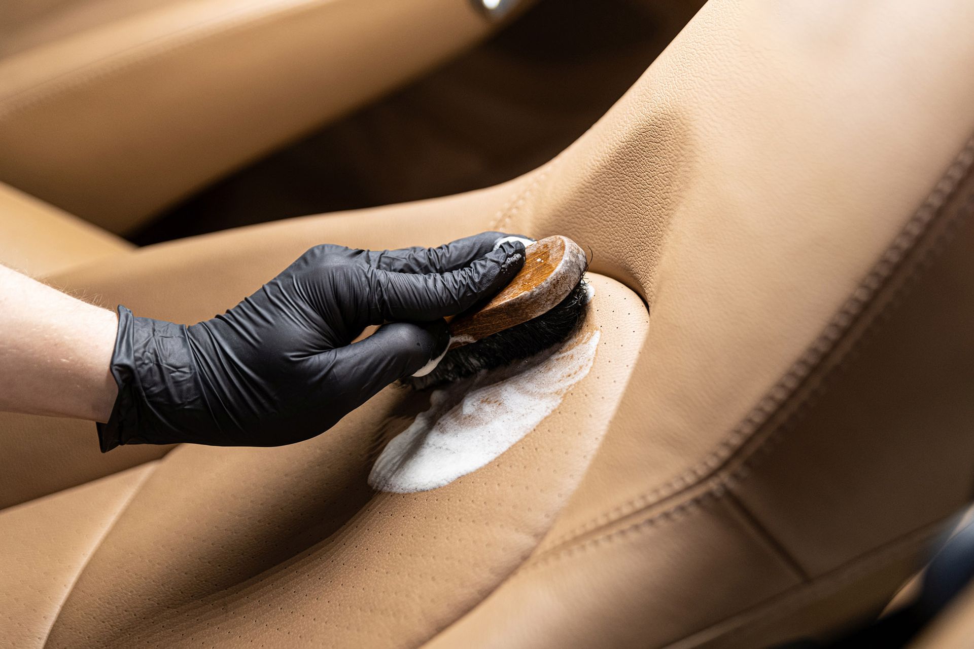 A gloved hand uses a brush and cleaning foam to scrub a tan leather car seat.