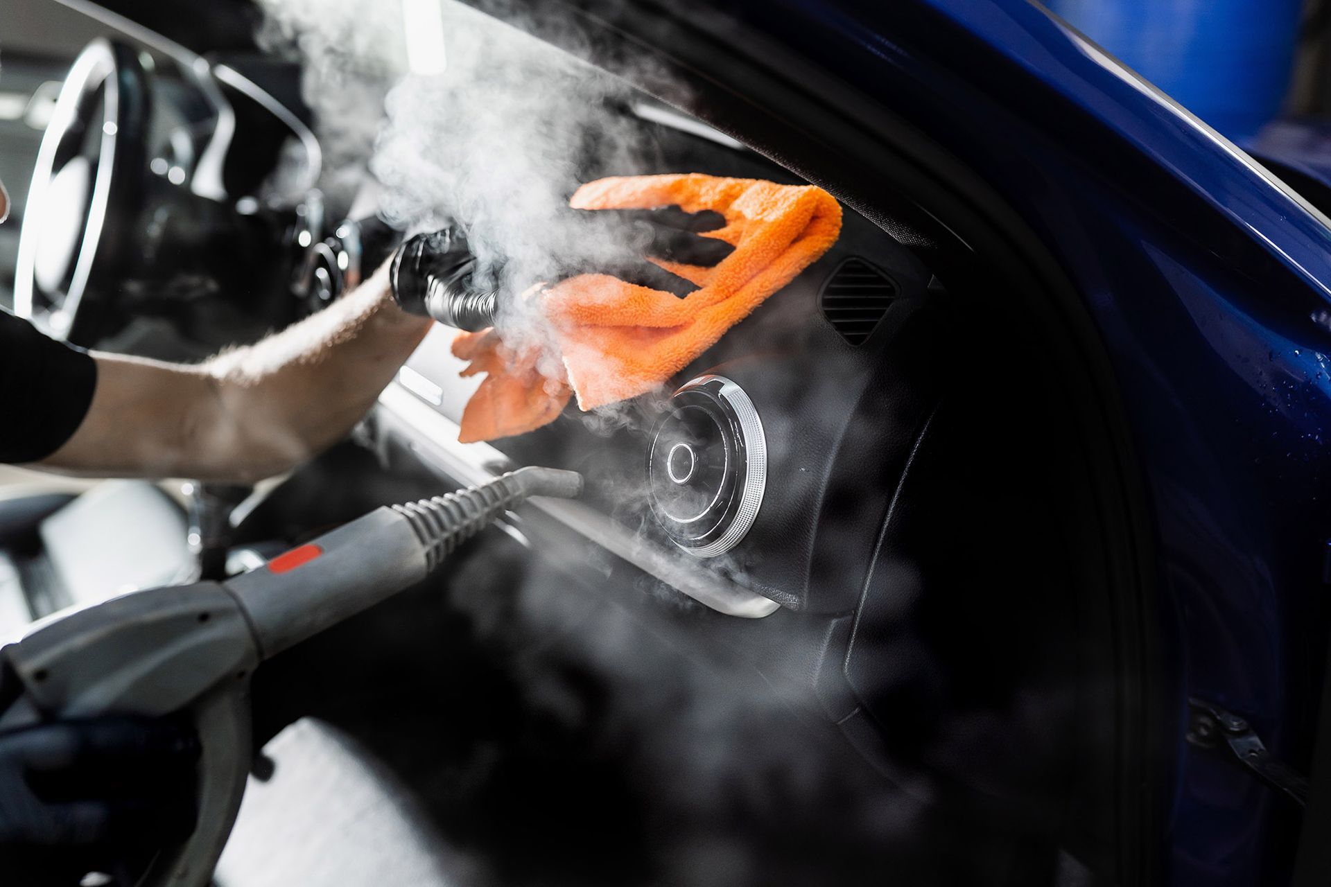 A professional detailer uses a steam cleaner and an orange microfiber cloth to clean a car's dashboard and air vent.