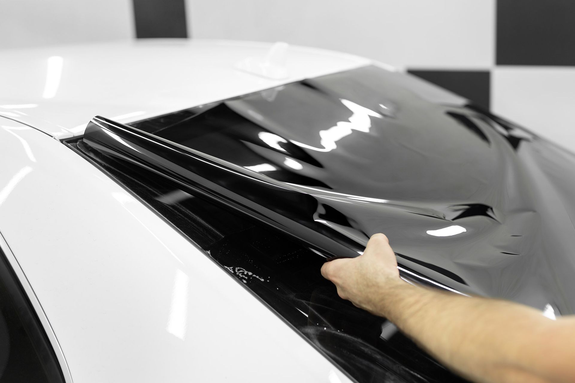 A person applying black window tint film to the rear windshield of a white car.