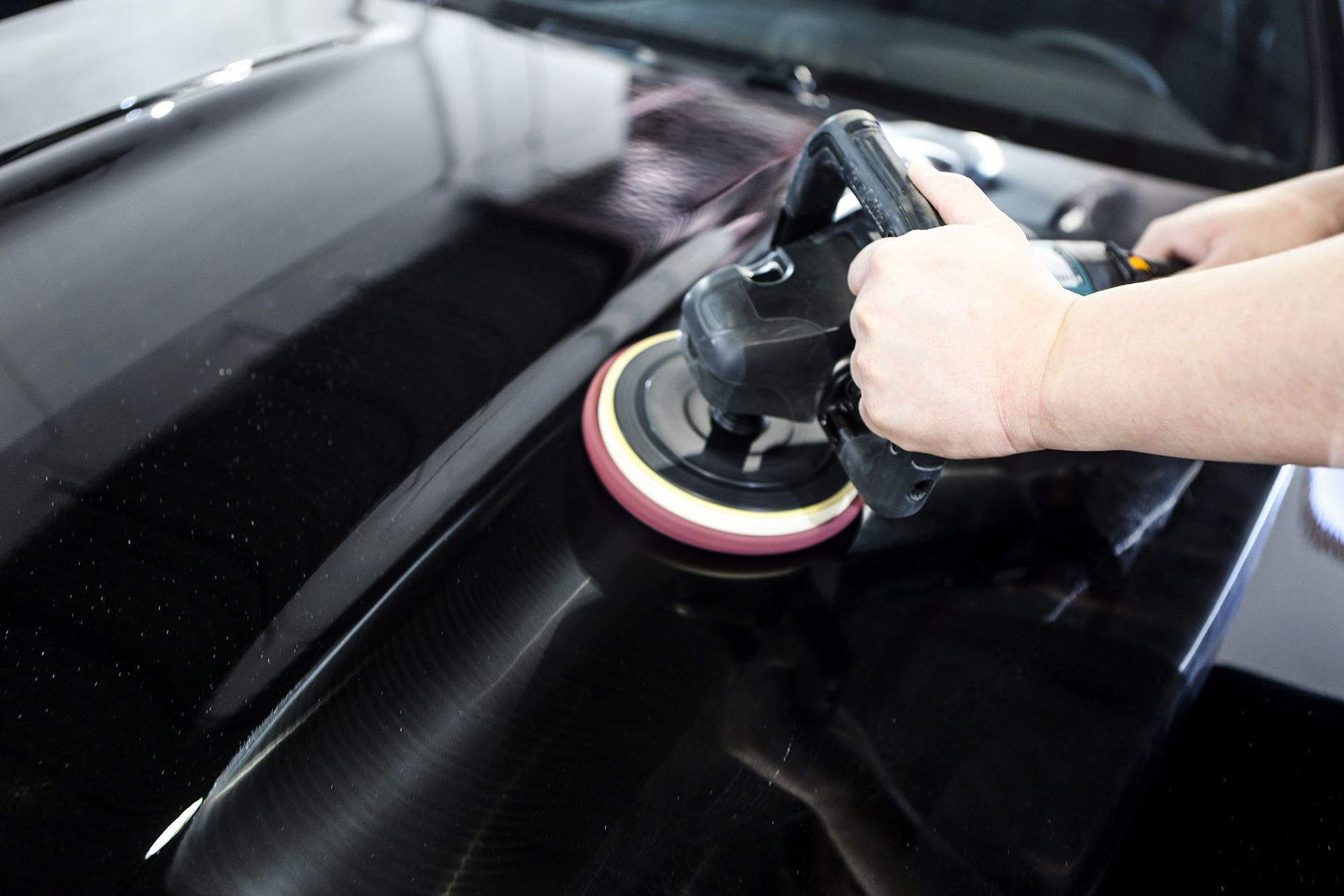 A person uses an electric buffer to polish the shiny black surface of a car.