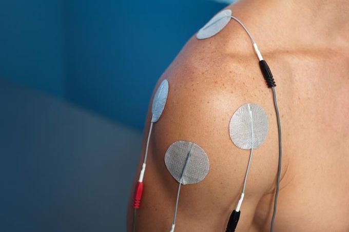 Shoulder with several electrode pads attached, wires connected; blue background.