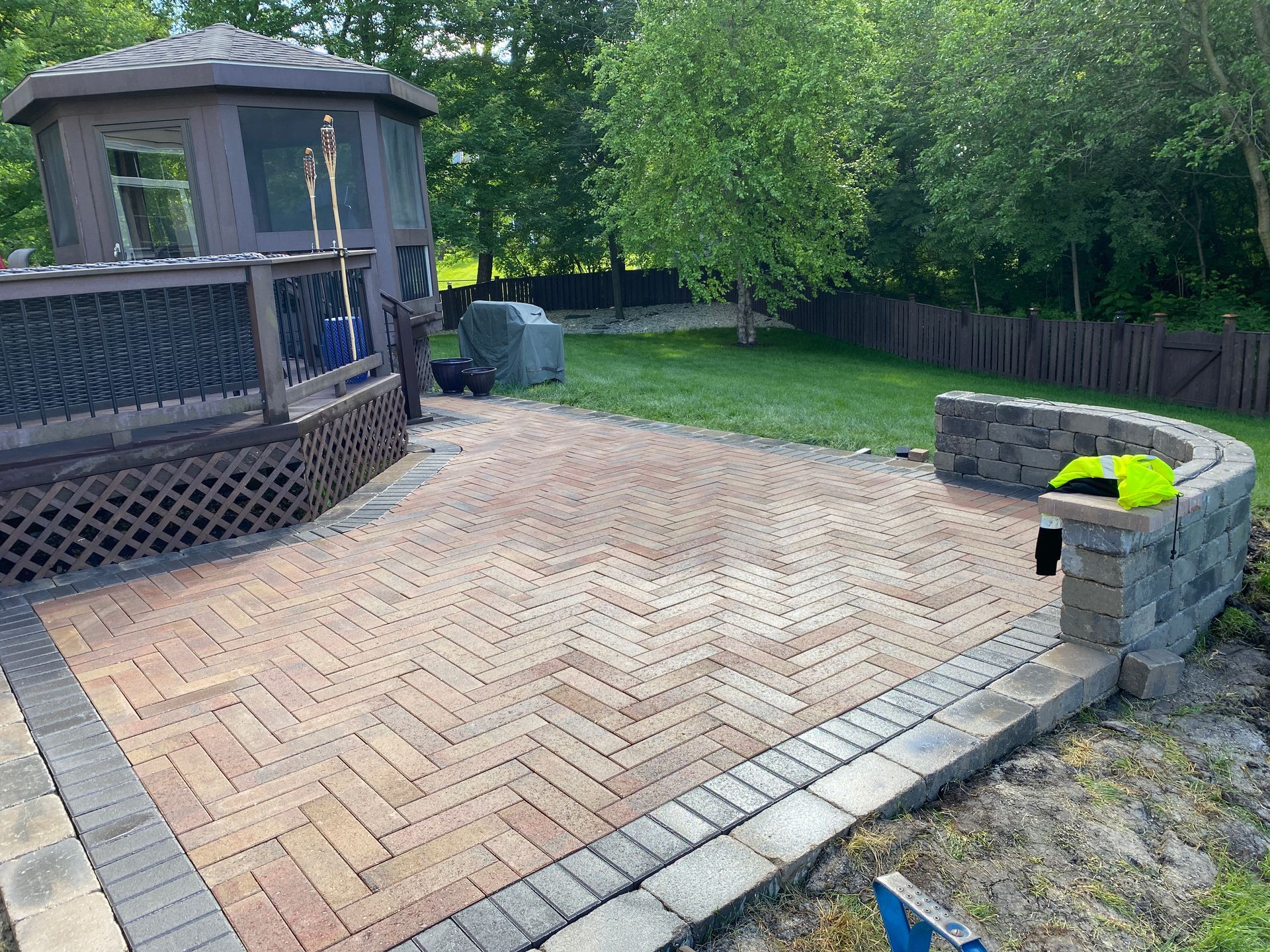Backyard patio with brick pavers, gazebo, raised stone fire pit, and green lawn bordered by trees