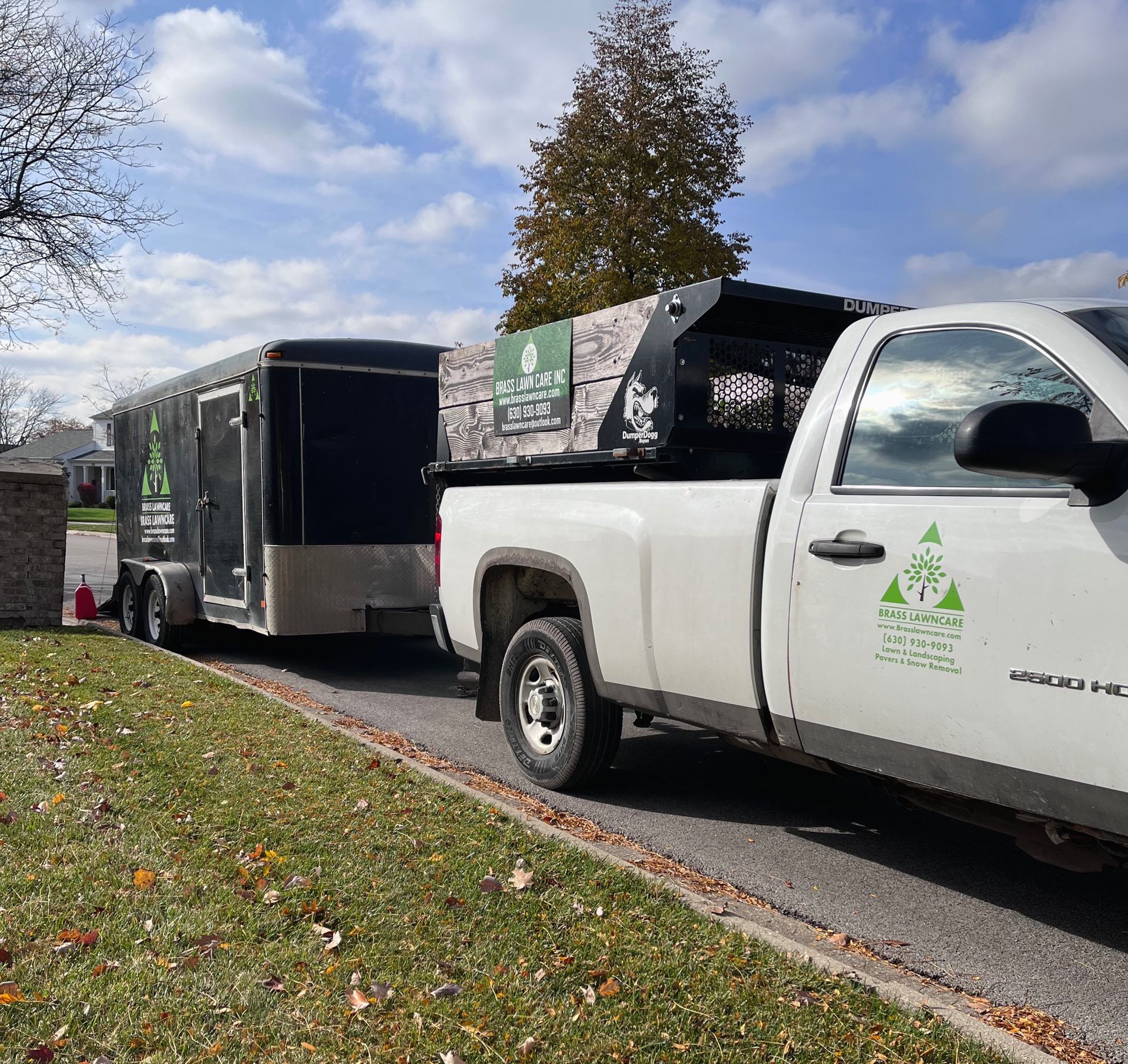 White utility truck towing a black enclosed trailer on a leafy street under a cloudy sky