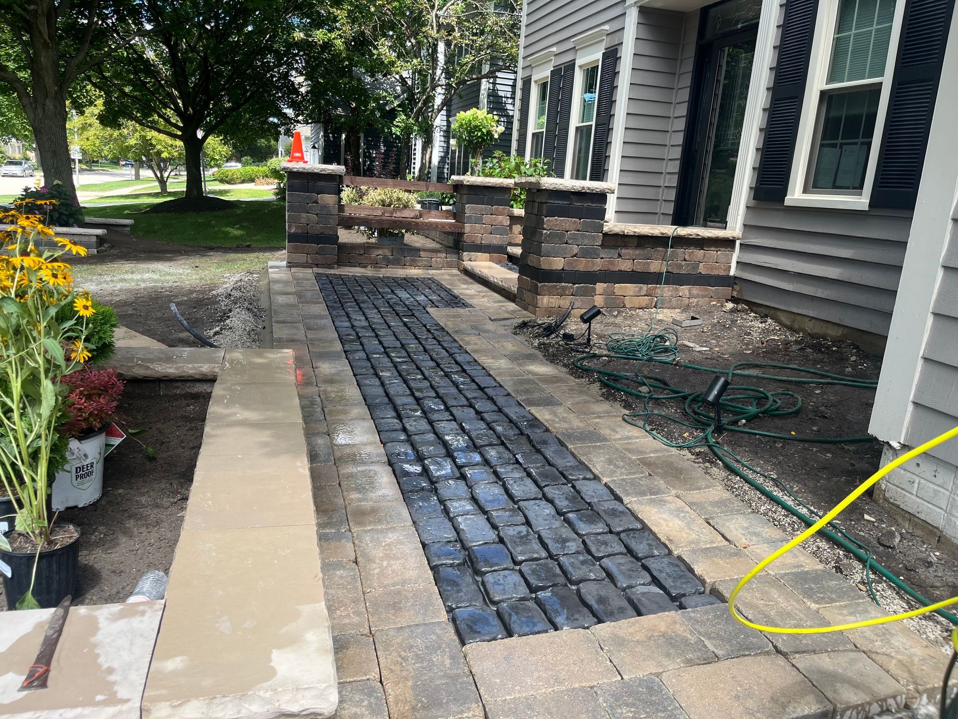 Front walkway under construction beside a house, with dark pavers, concrete edging, and garden beds.