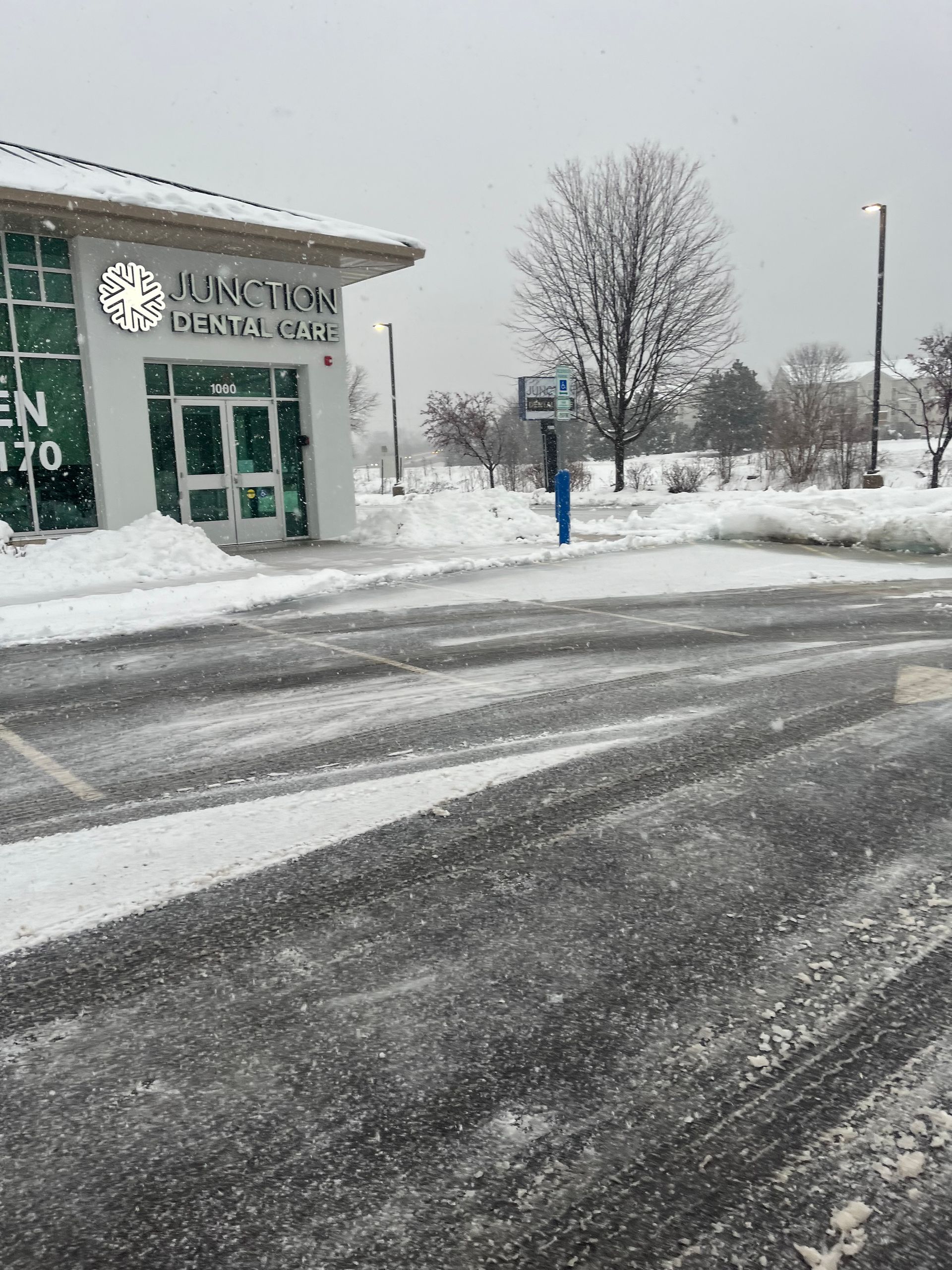 Snowy parking lot outside a building with a green door and a leafless tree.