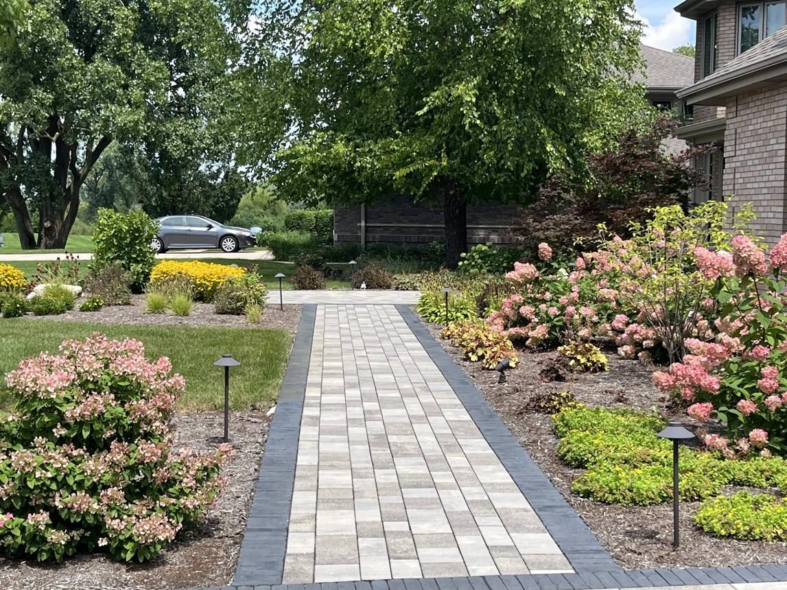 Stone walkway through a landscaped garden with pink flowers and trees leading to a house