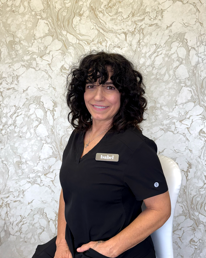 Woman with dark curly hair in black scrubs smiles near a window.