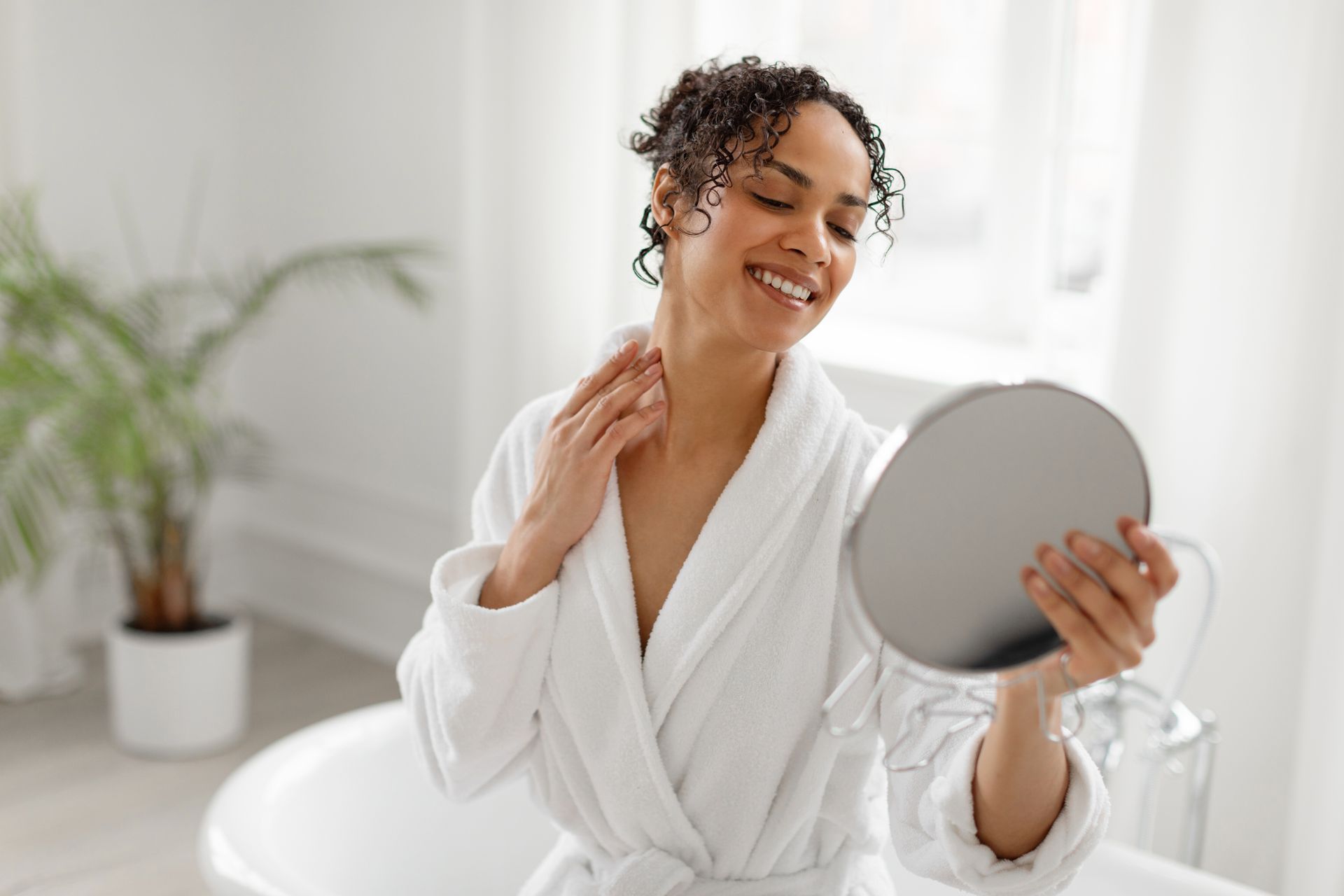 Woman in a white robe applying skincare while smiling at a round mirror in a bright room