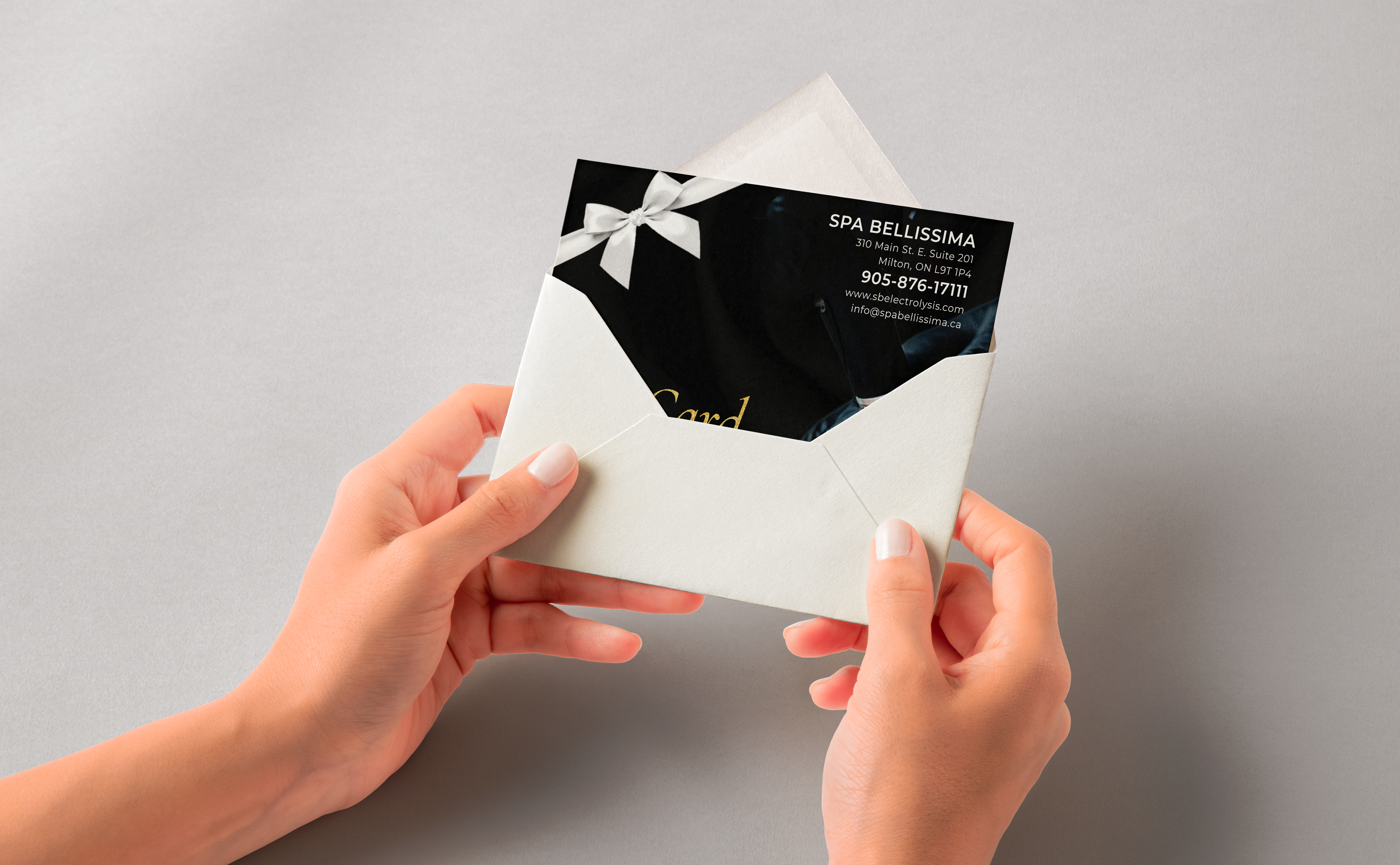 Hands holding a white envelope-style card with a black insert and small white bow on a gray background