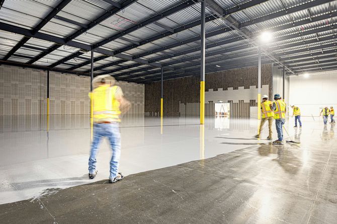 Workers in yellow high-visibility vests apply a smooth gray epoxy coating to a large, unfinished warehouse concrete floor.