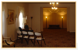 Interior of a funeral home chapel with chairs, a window, and a lit stage area.