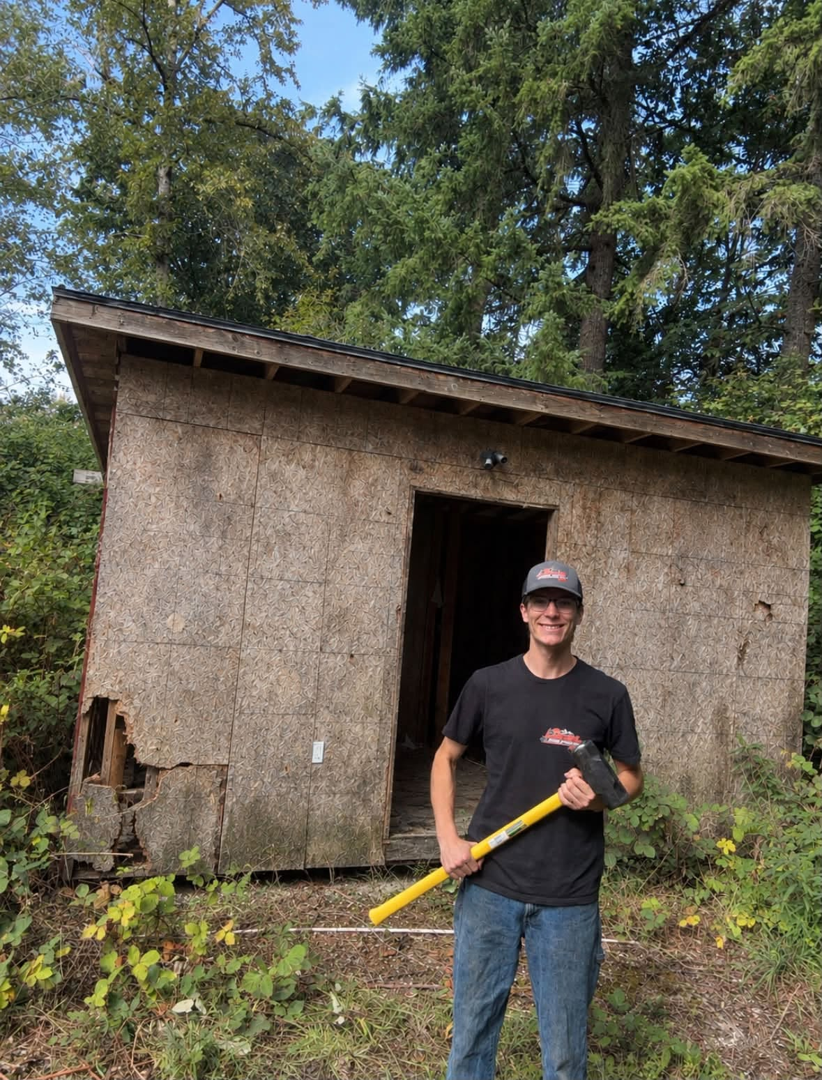 Person in a black shirt and cap holding a yellow tool in front of a small weathered shed in a wooded area