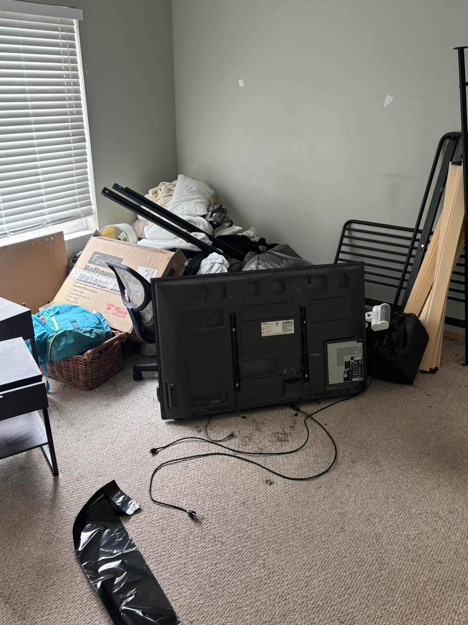 Cluttered room with a fallen black TV stand, guitar, boxes, and scattered items near a window with blinds