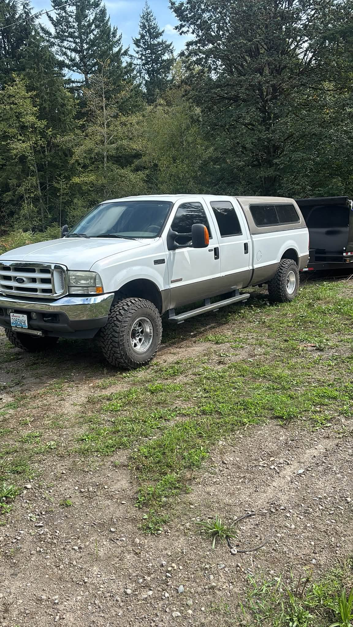 White pickup truck parked on a gravel driveway beside trees and a dark vehicle in the background