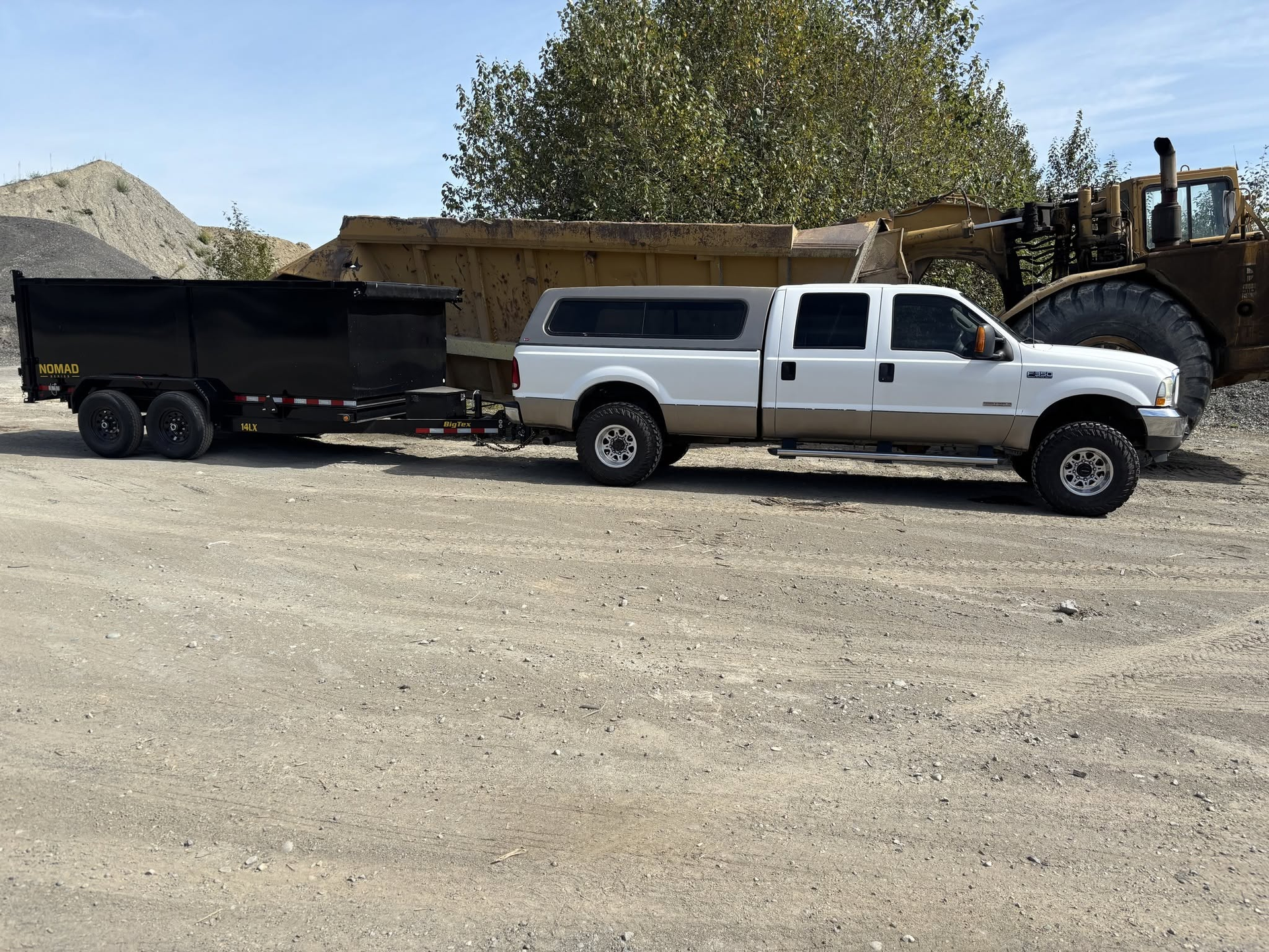 White pickup truck towing a black dump trailer on a gravel lot near construction equipment
