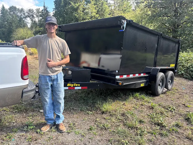 Man standing beside a black dual-axle utility trailer hitched to a white truck in a grassy area