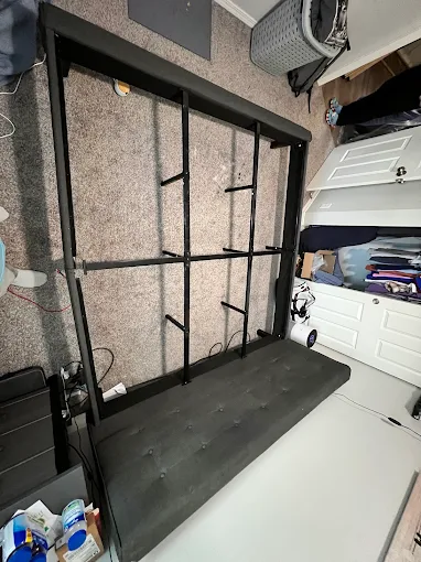 Black metal bed frame on its side in a carpeted room beside a mattress and white drawers.