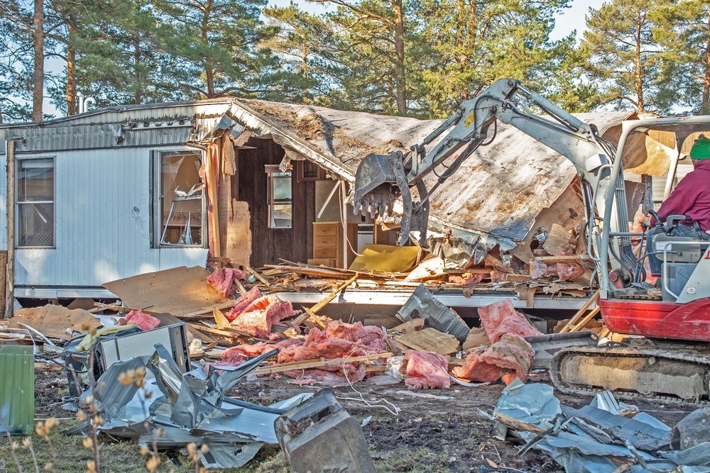 Damaged home with collapsed roof and debris, with an excavator clearing rubble in the yard