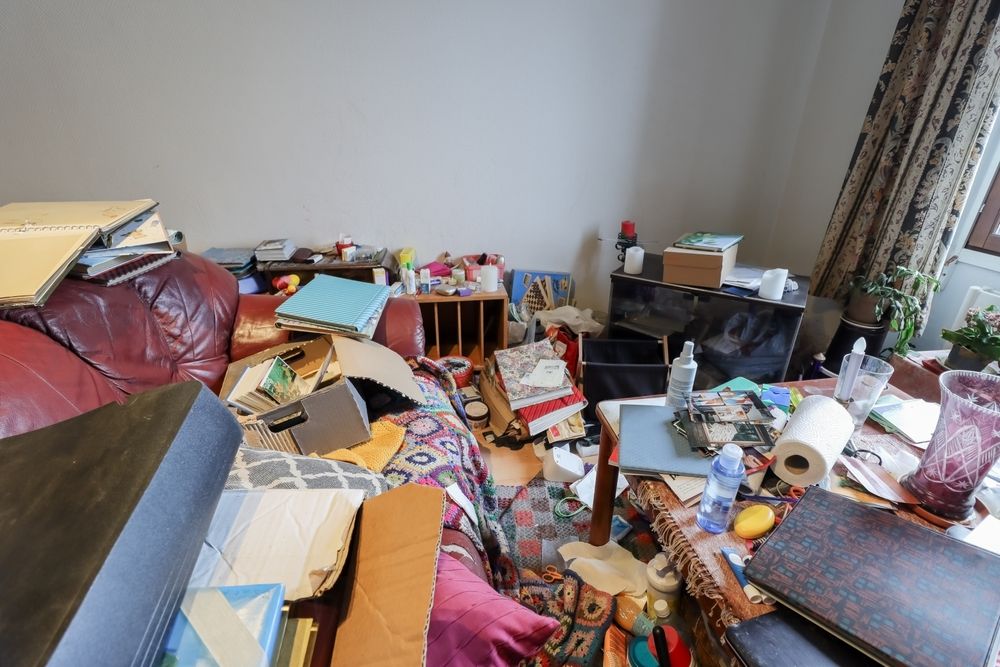 Cluttered living room with scattered toys, papers, and household items across the floor and furniture