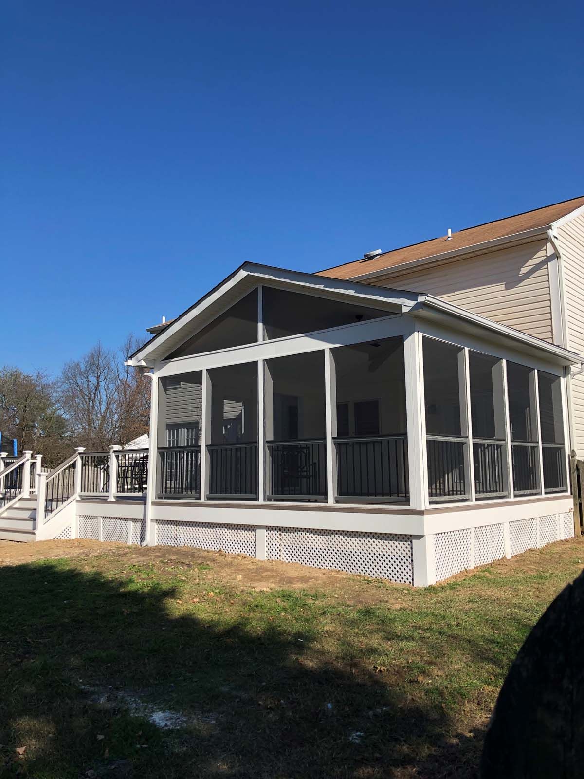 Screened-in porch addition with dark railing, white trim, and a deck, attached to a two-story house on a sunny day.