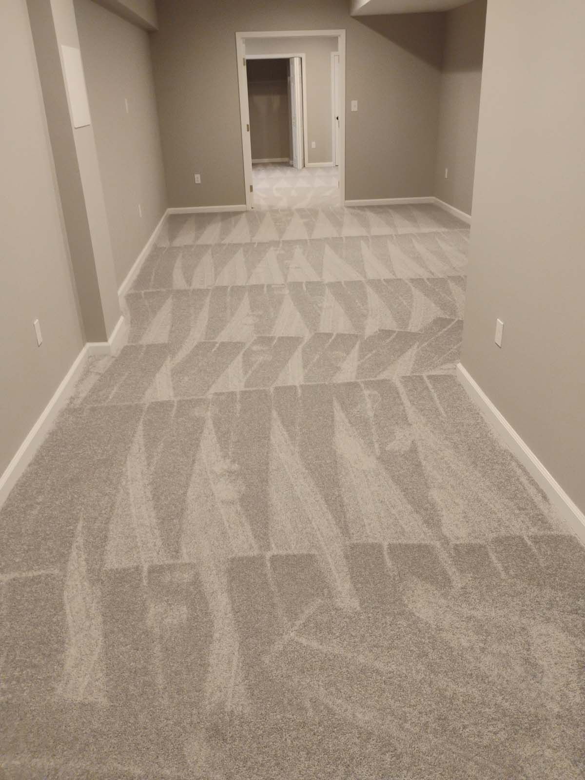 A long, carpeted room with a repeating geometric pattern in shades of gray, leading to an open doorway.