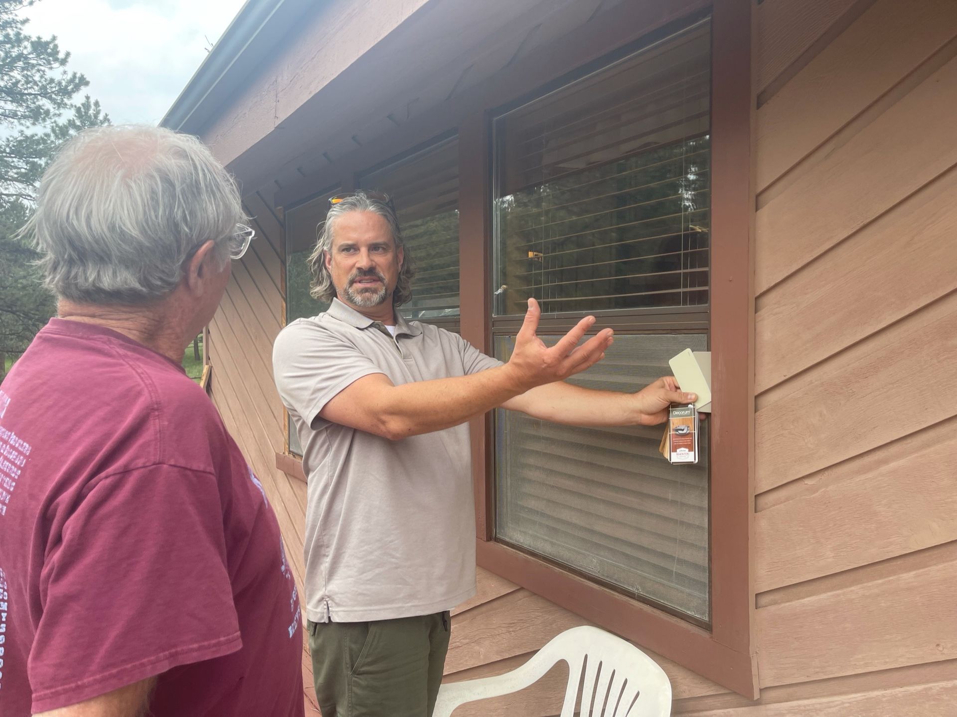 Man explaining something next to a window to another man; brown exterior, sunny day.