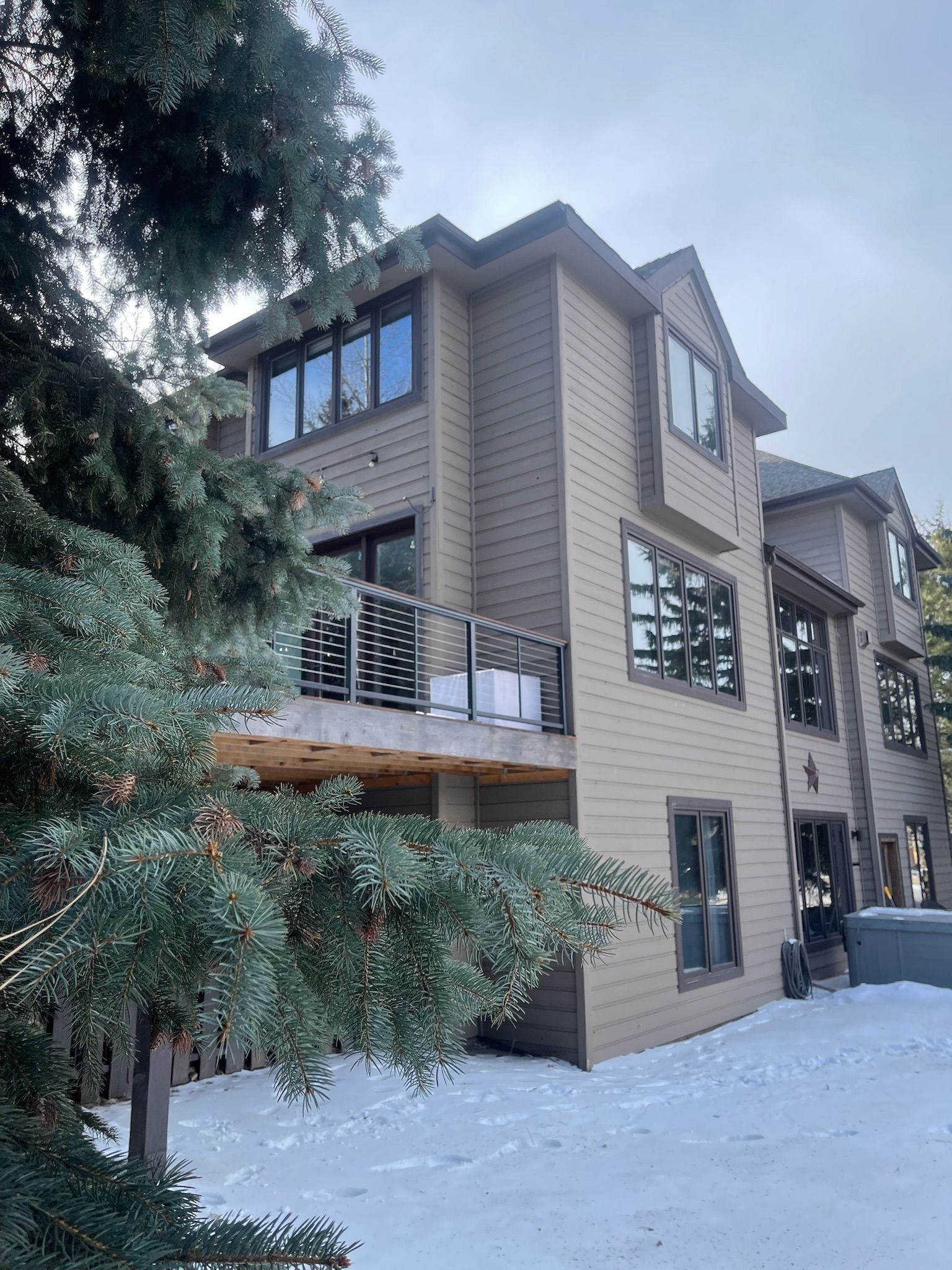 Multi-story beige house with snow-covered ground, wooden deck, and surrounding trees.