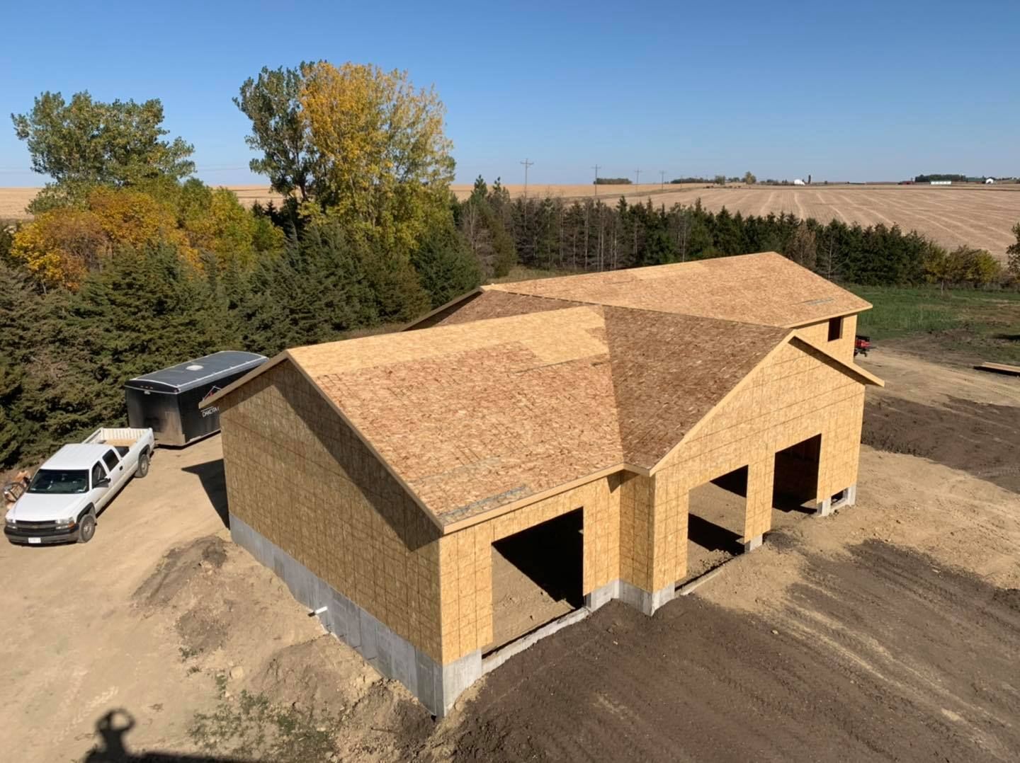 Aerial view of a partly built wooden house with a shingled roof beside parked trucks and farmland.