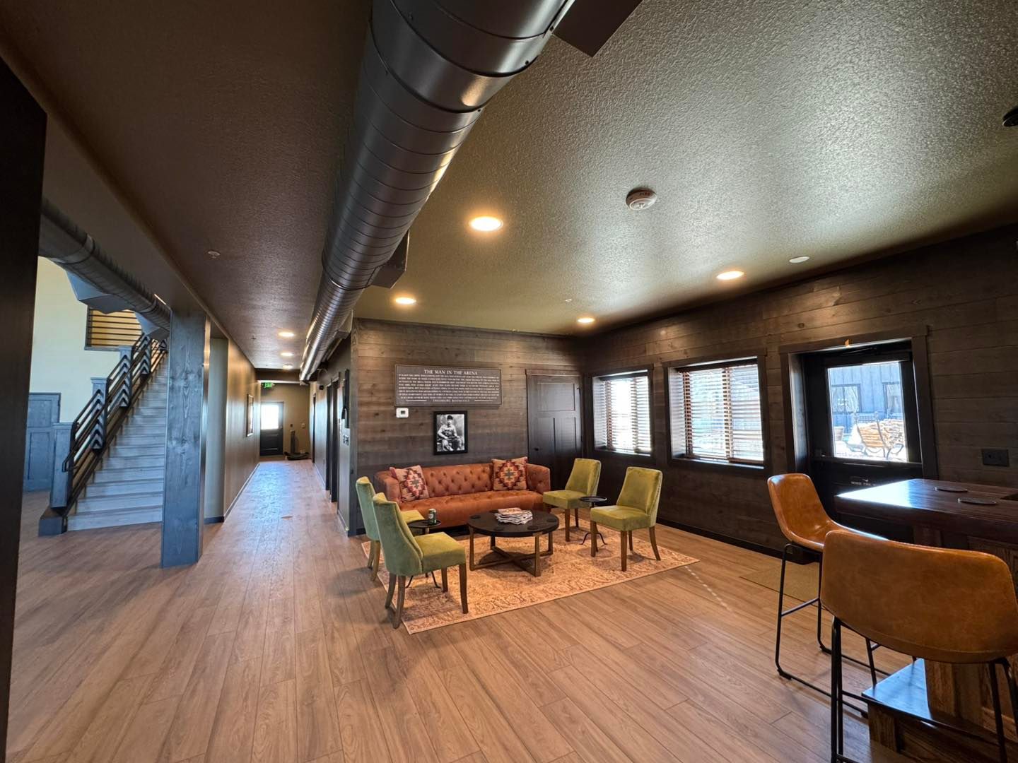 Modern lounge with brick walls, wood floors, round table, chairs, stools, and windows along one side.