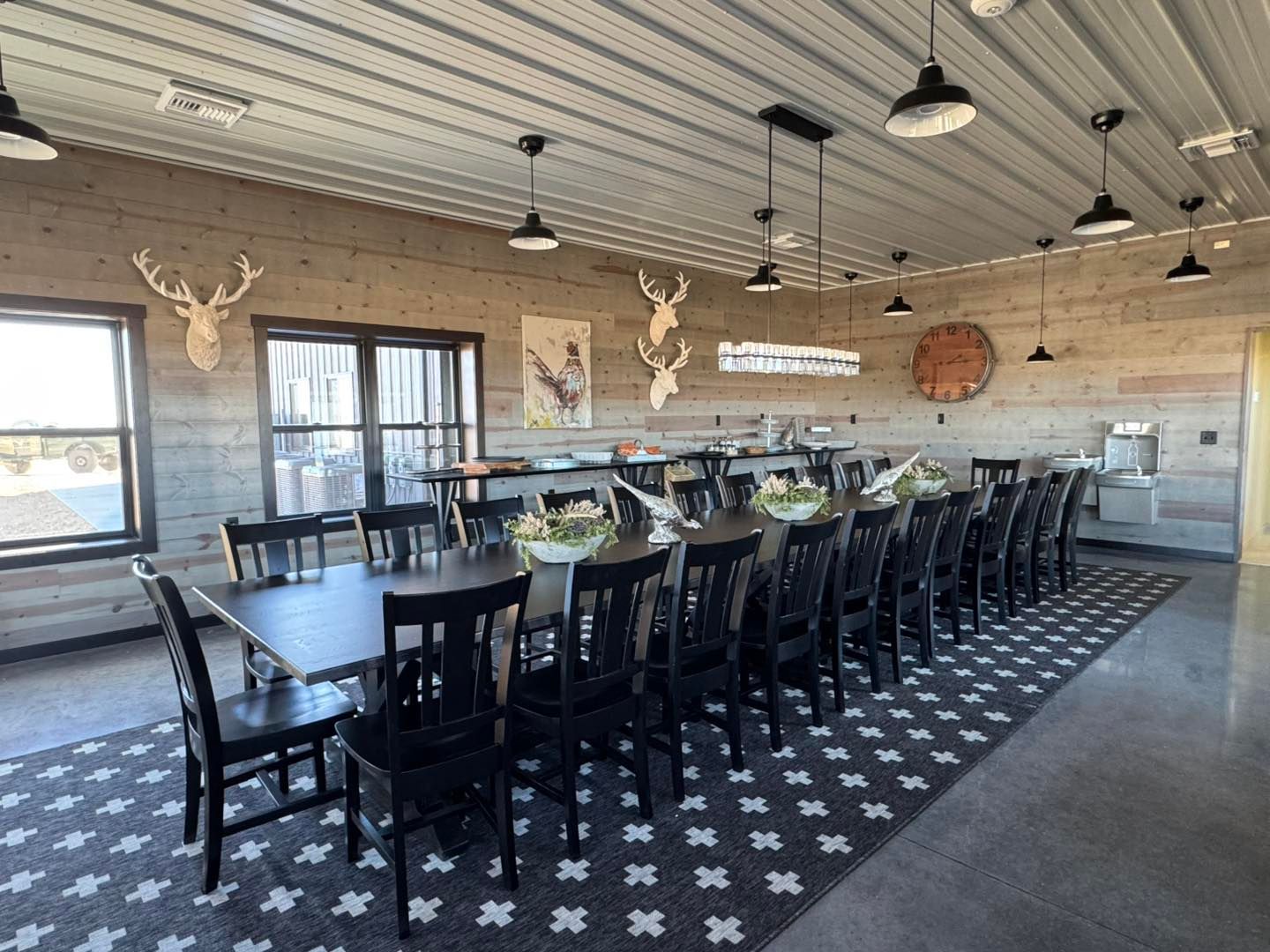 Spacious rustic dining hall with long tables, black chairs, hanging lights, and deer wall decor