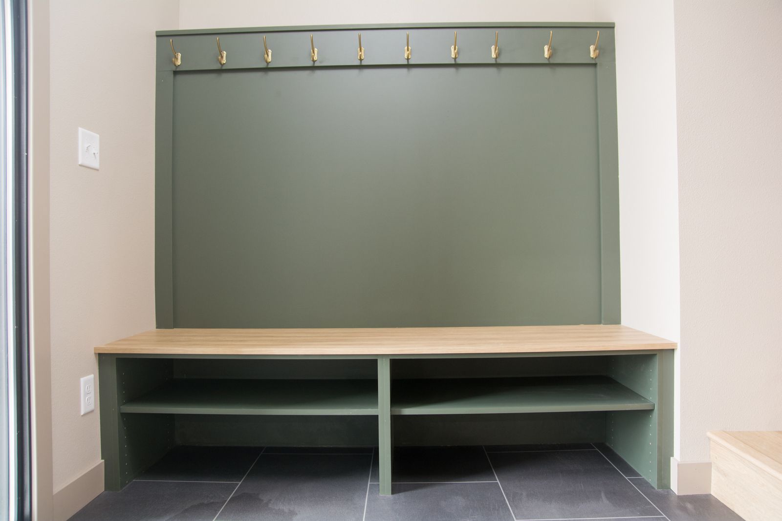 Mudroom bench with beige seat, olive-green wall panel, and hooks above, on dark tile floor