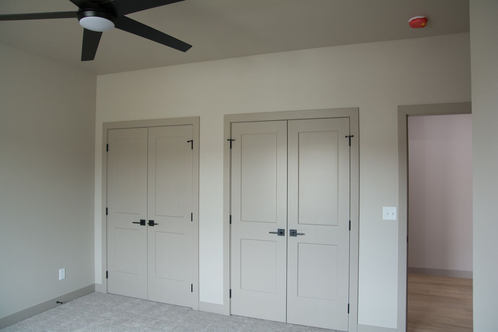 Empty bedroom with beige carpet, white double closets, ceiling fan, and an open doorway to another room