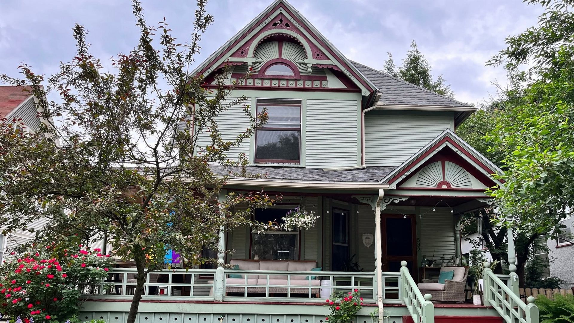Victorian-style house with white trim and a front porch, surrounded by trees and greenery.