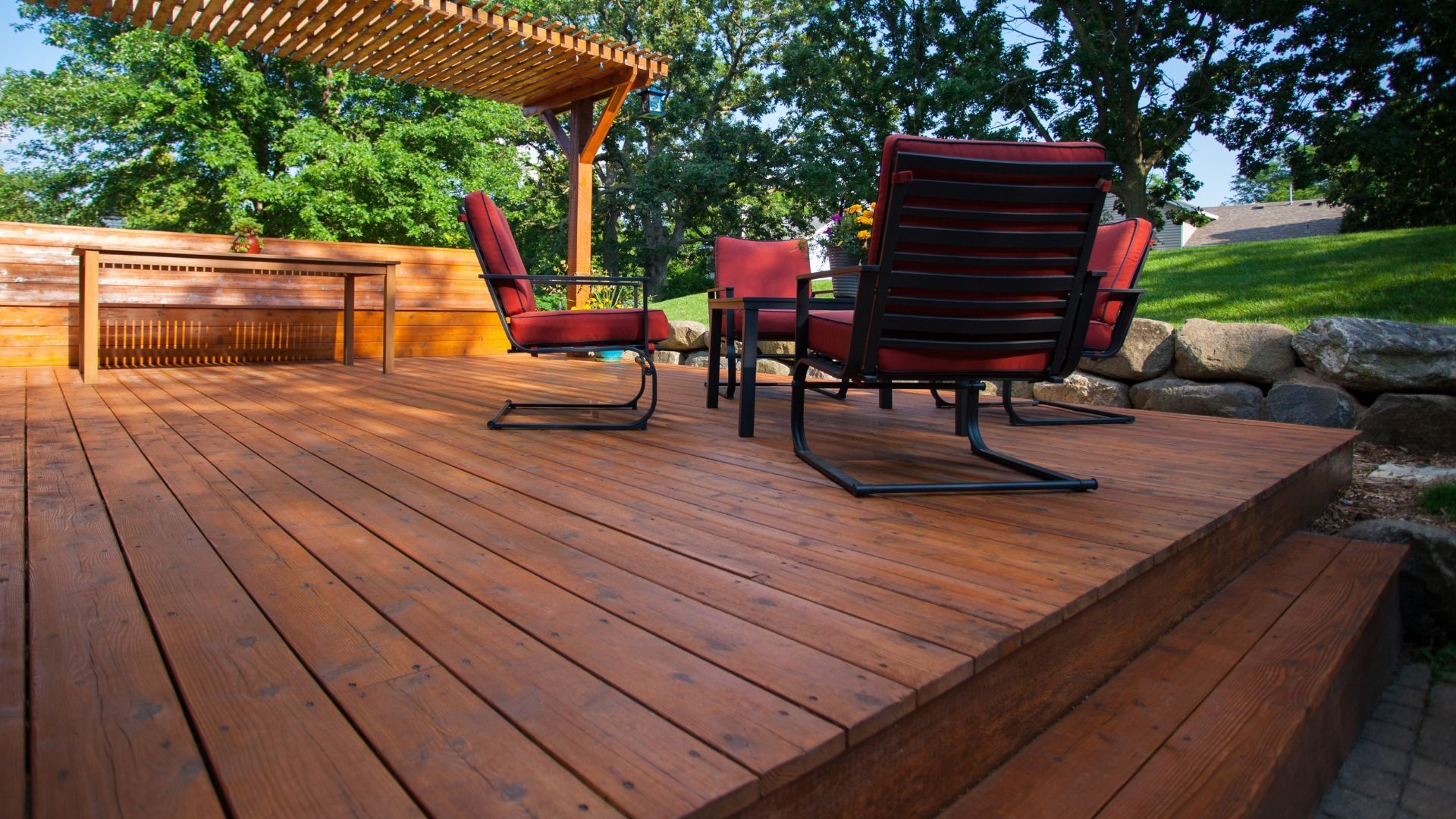 Wooden deck patio with red cushioned chairs under a pergola in a sunny backyard