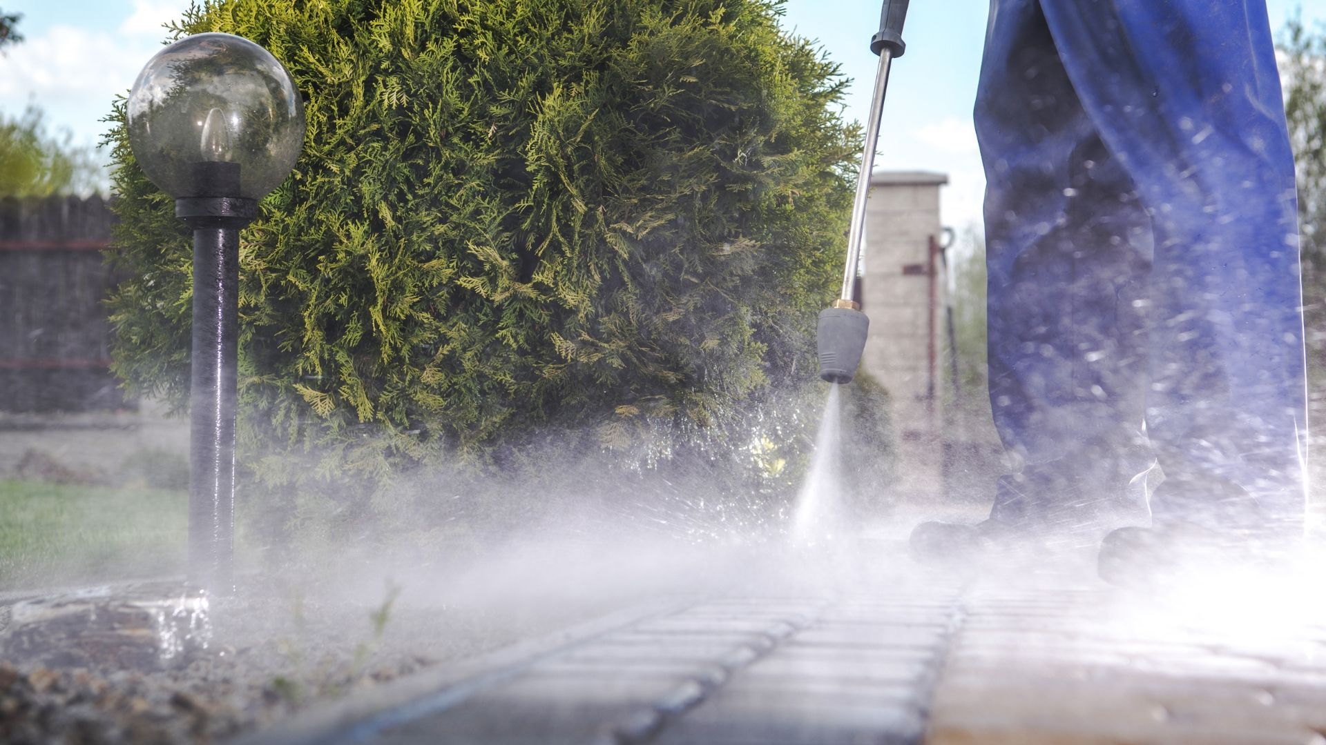Person pressure-washing a patio, with water spray, a green bush, and a lamp post in the background