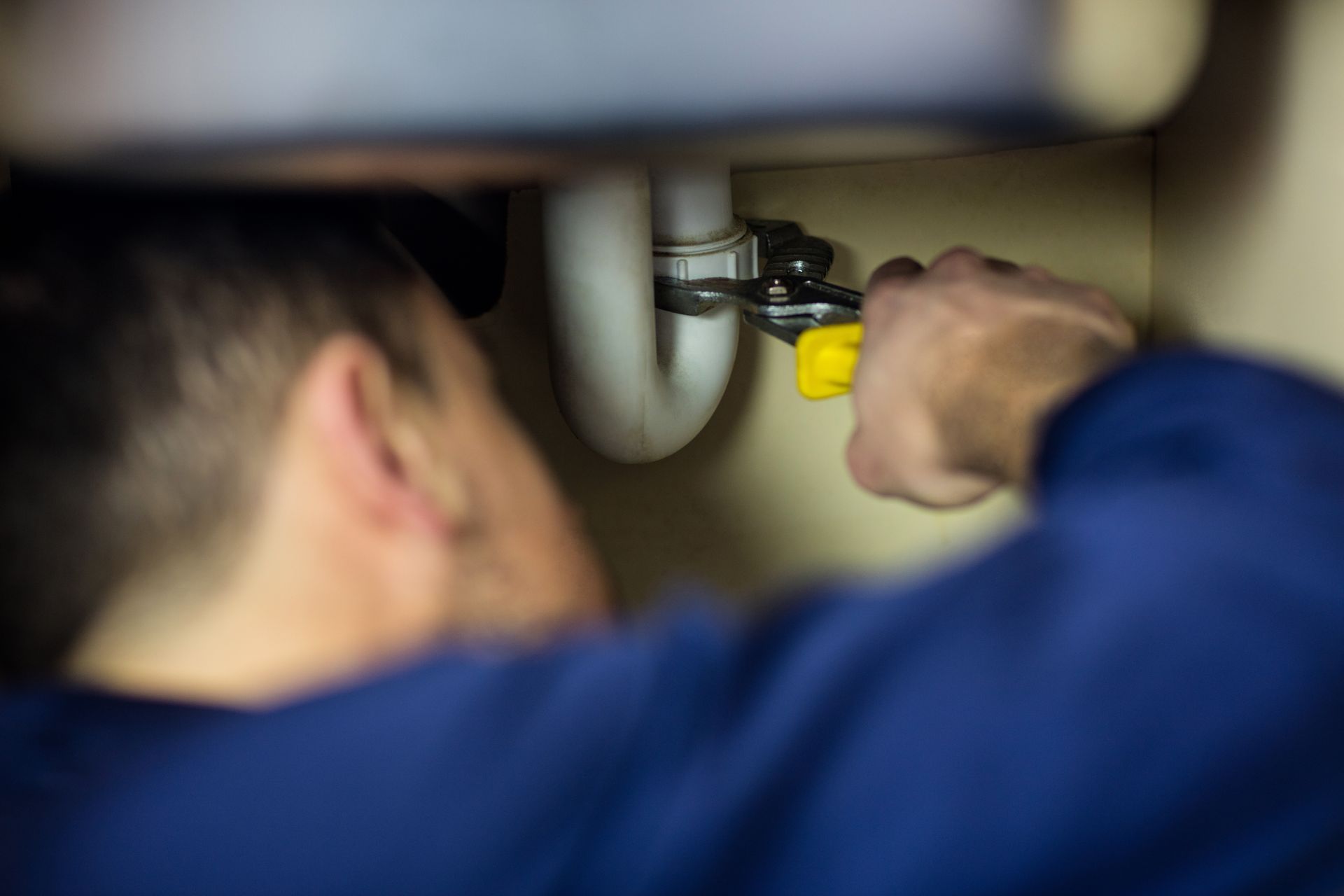 A plumber in a blue shirt tightens a PVC pipe under the sink.
