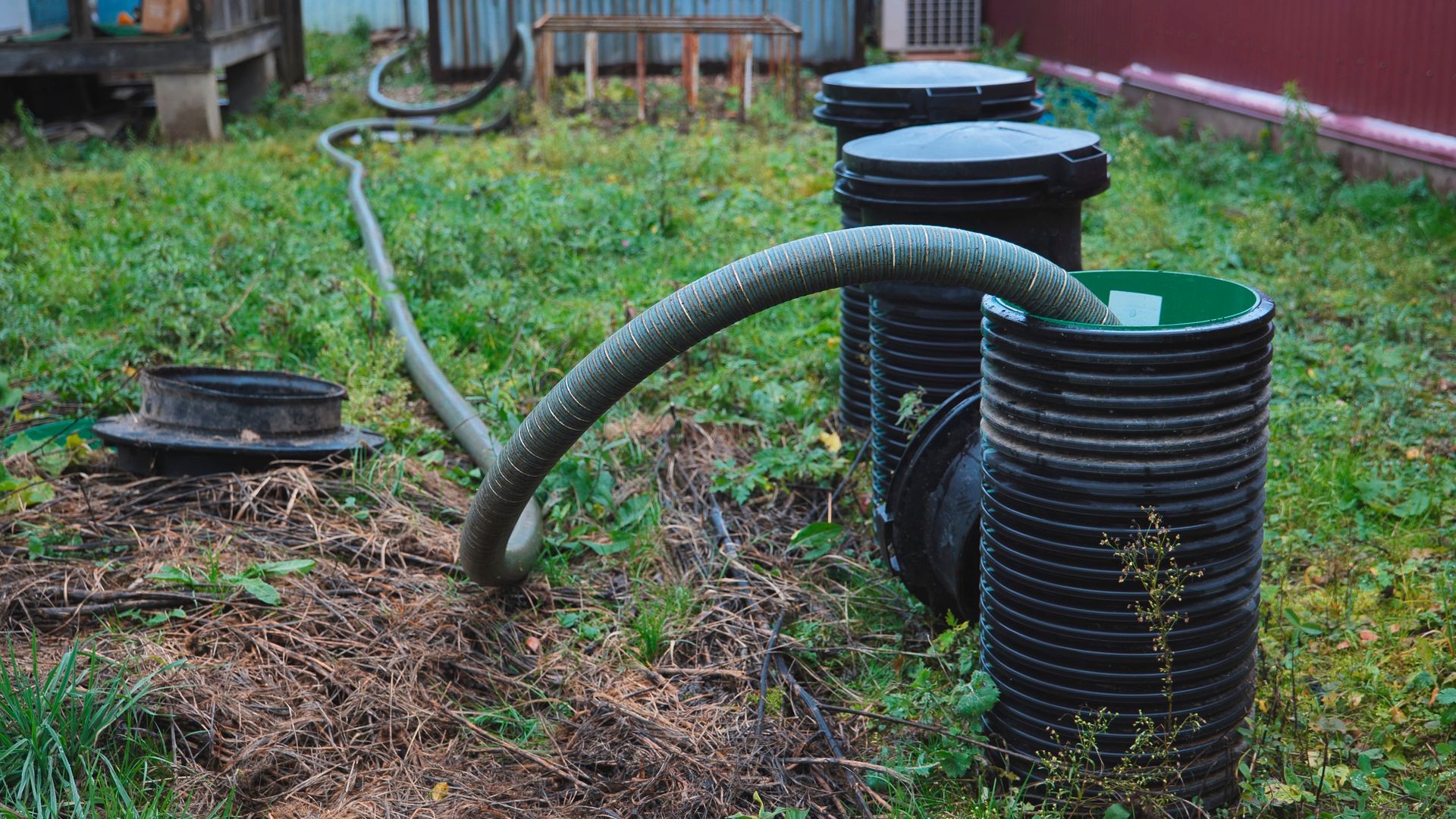 Garden hoses and black corrugated pipes on damp grass beside a yard pump and a wooden fence. Garden hoses and black corrugated pipes on damp grass beside a yard pump and a wooden fence.