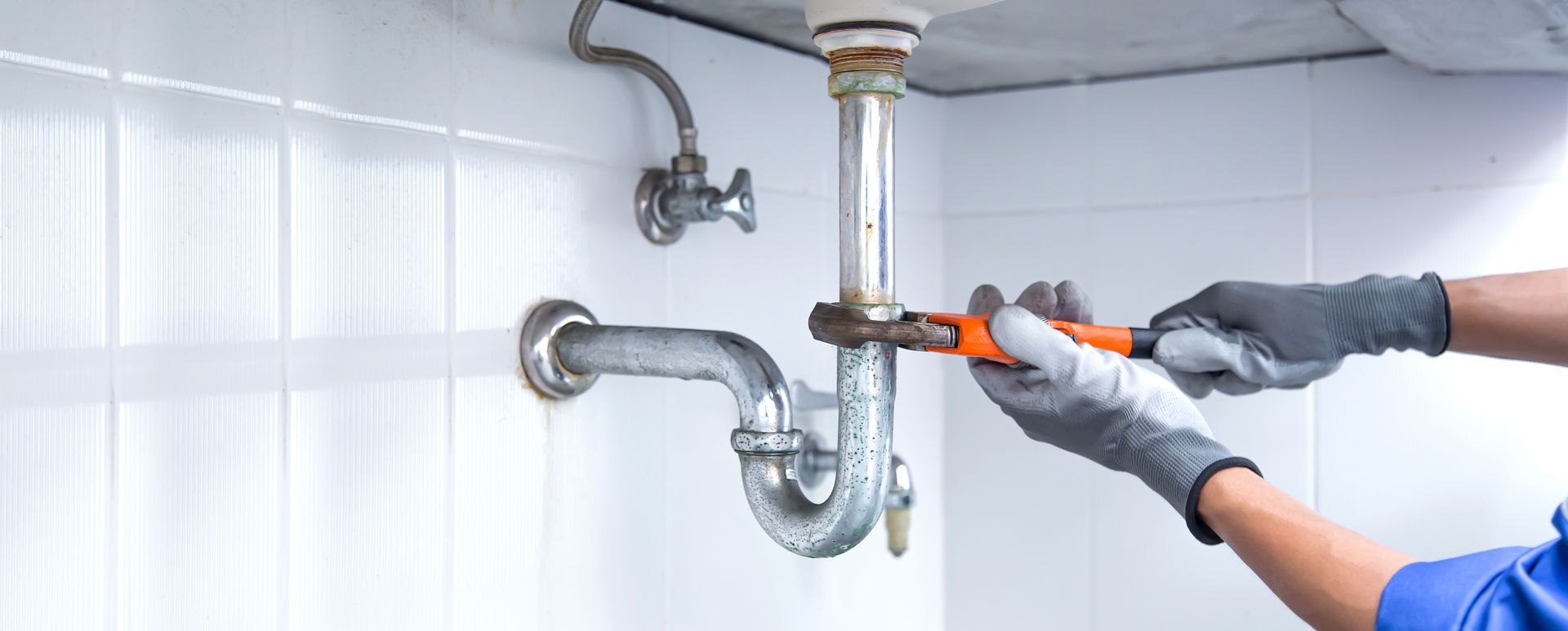 Close-up of gloved hands tightening a chrome P-trap fitting with an adjustable wrench under a sink.