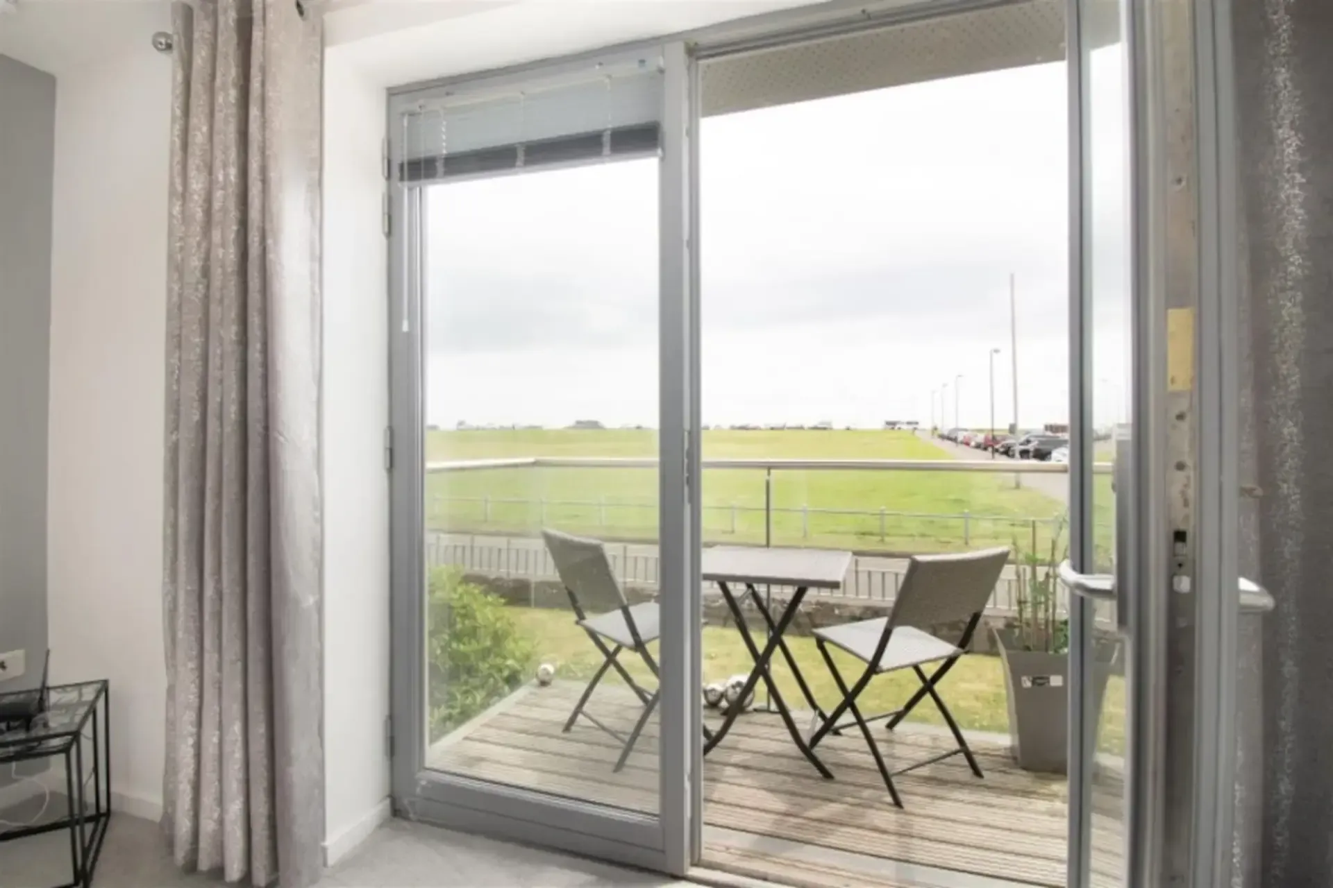 Sliding glass doors open to a patio with table and chairs overlooking a green field and cloudy sky