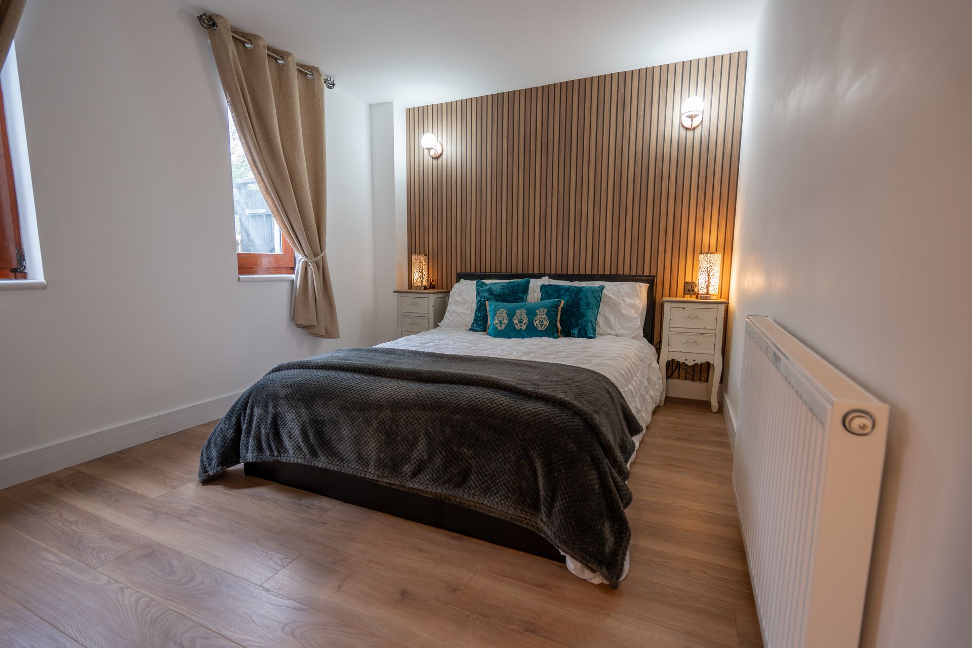Bright bedroom with a double bed, wood-paneled headboard, curtains, and two small windows.