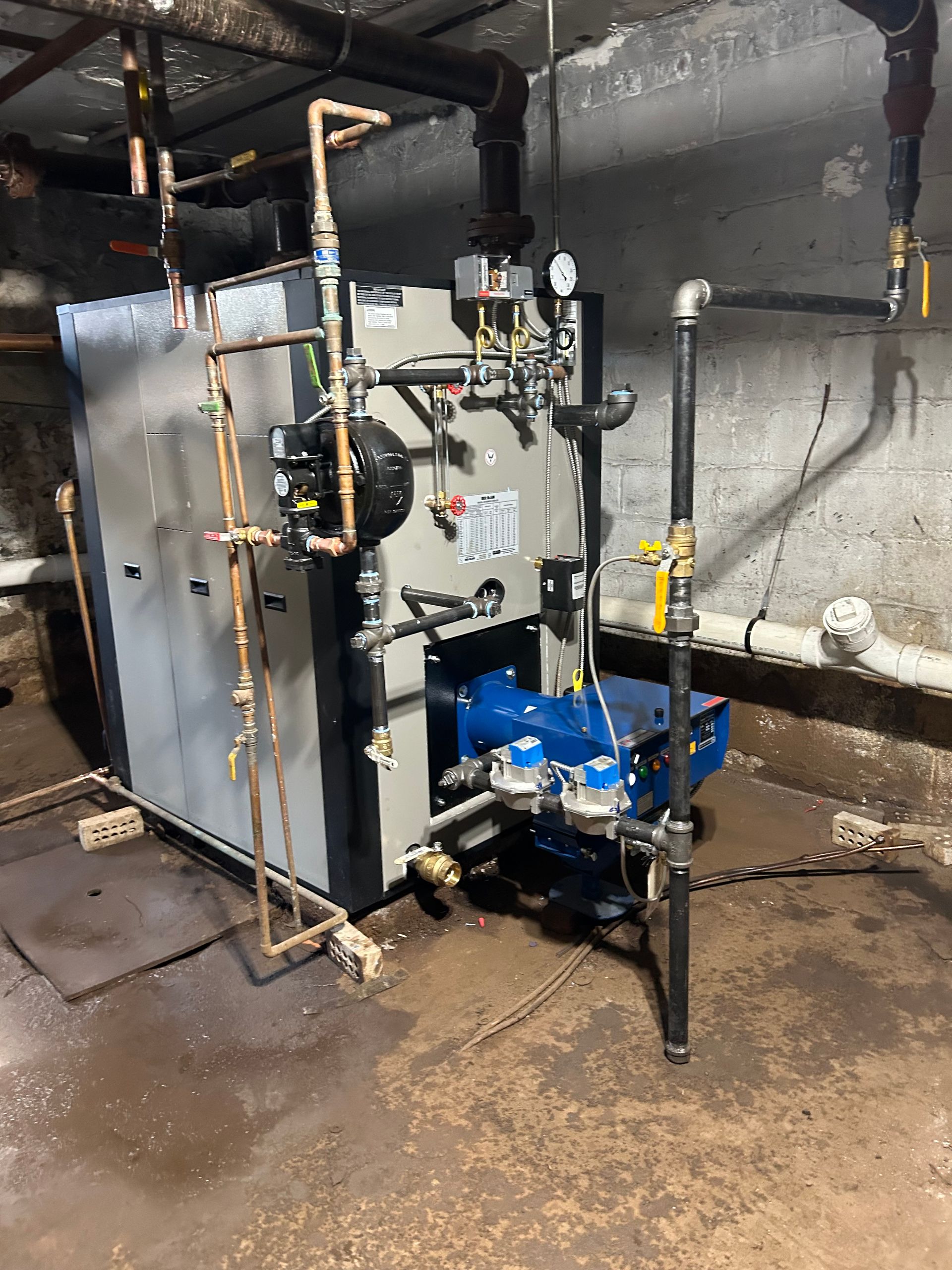 A boiler in a basement, with copper pipes and a blue burner. Grey brick wall background.
