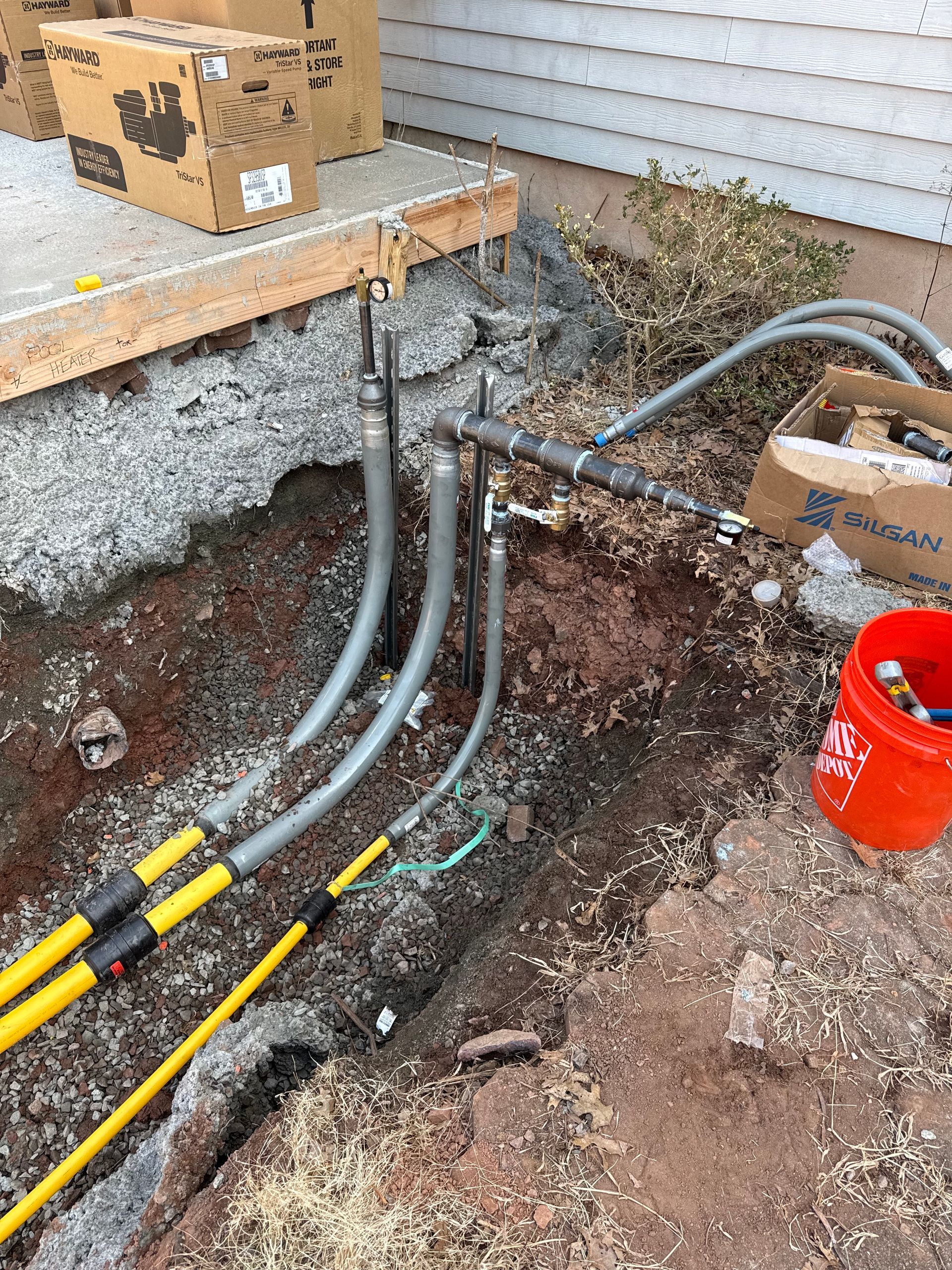 Electrical wires in trench, connecting to a house; includes conduit, yellow cables, and red bucket.