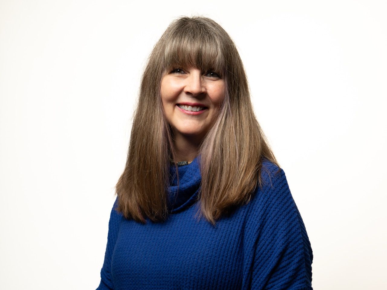 Woman with long, graying hair smiles, wearing a blue sweater. White background.