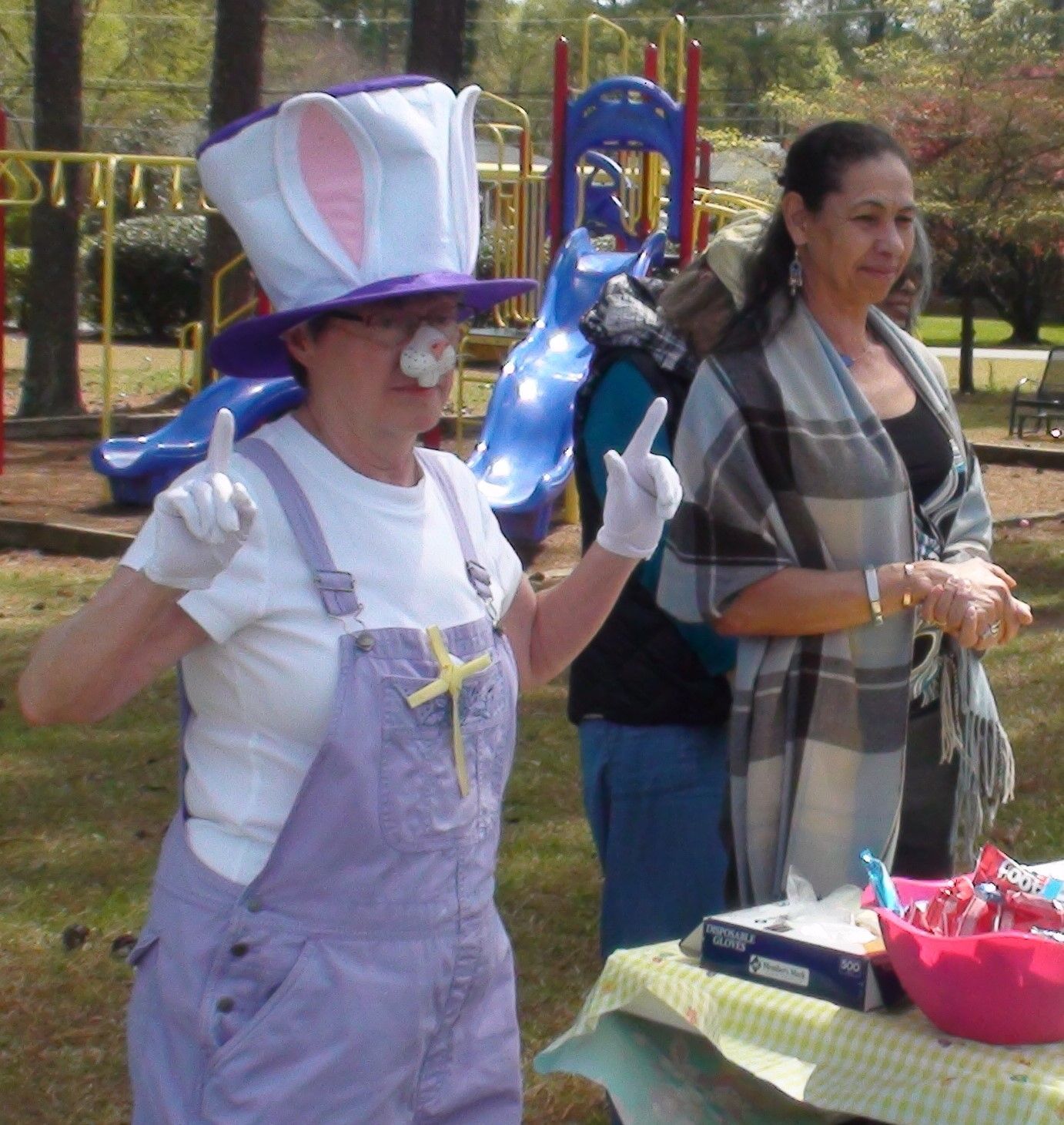 A woman wearing a bunny hat and overalls stands in front of a playground