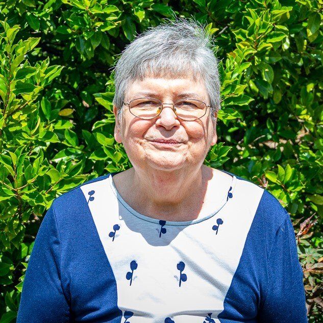 A woman wearing glasses and a blue and white shirt is standing in front of a bush.