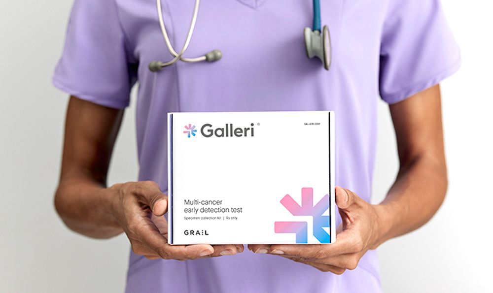 Nurse holding a Galleri cancer detection test box. The box is white with the Galleri logo and colorful design.